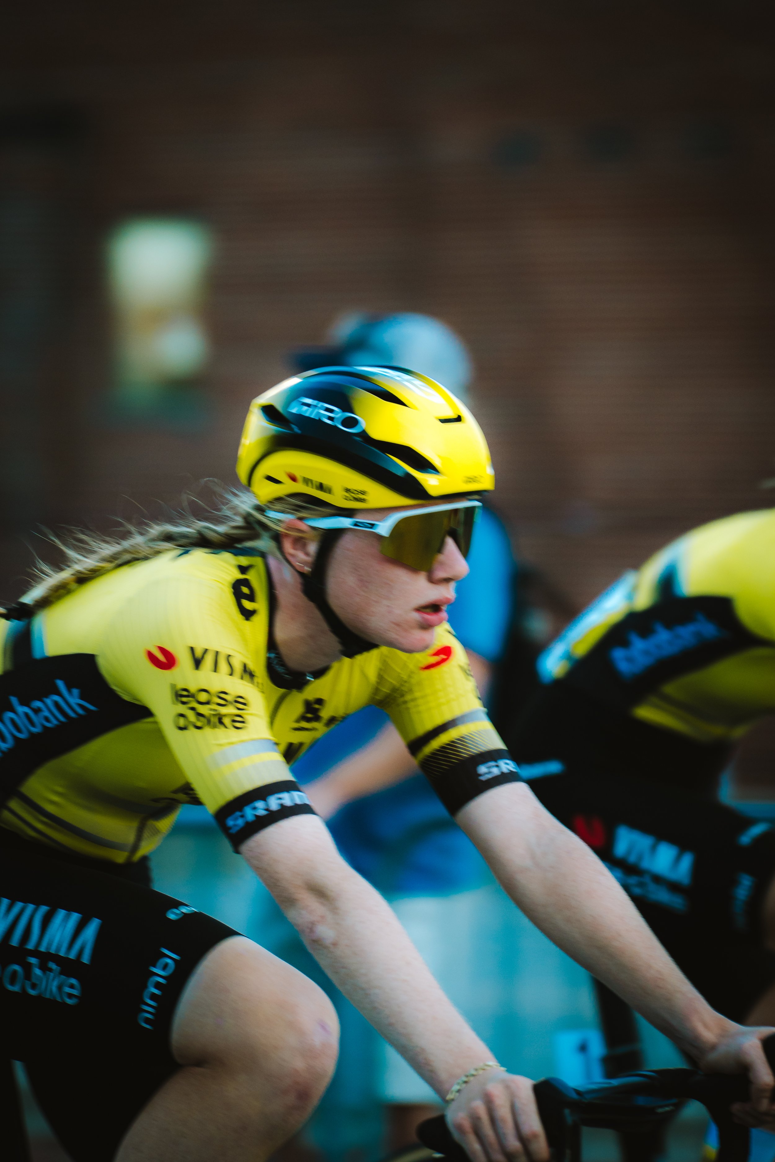 A female cyclist wearing a yellow team uniform, helmet, and sunglasses, riding in a race with a focused expression.