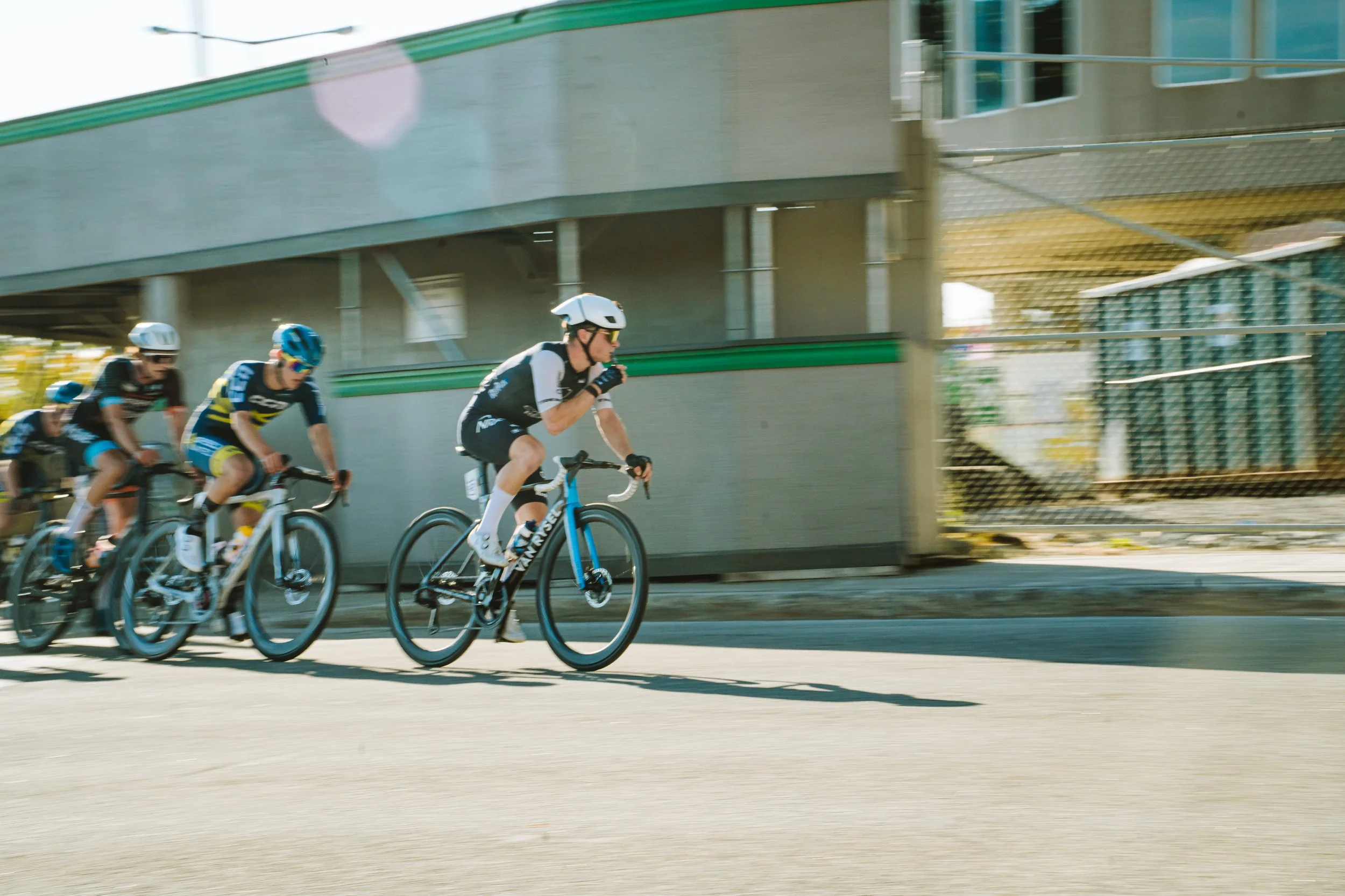 A group of competitive cyclists riding on a city street, with a bridge overhead and a fence on the side.