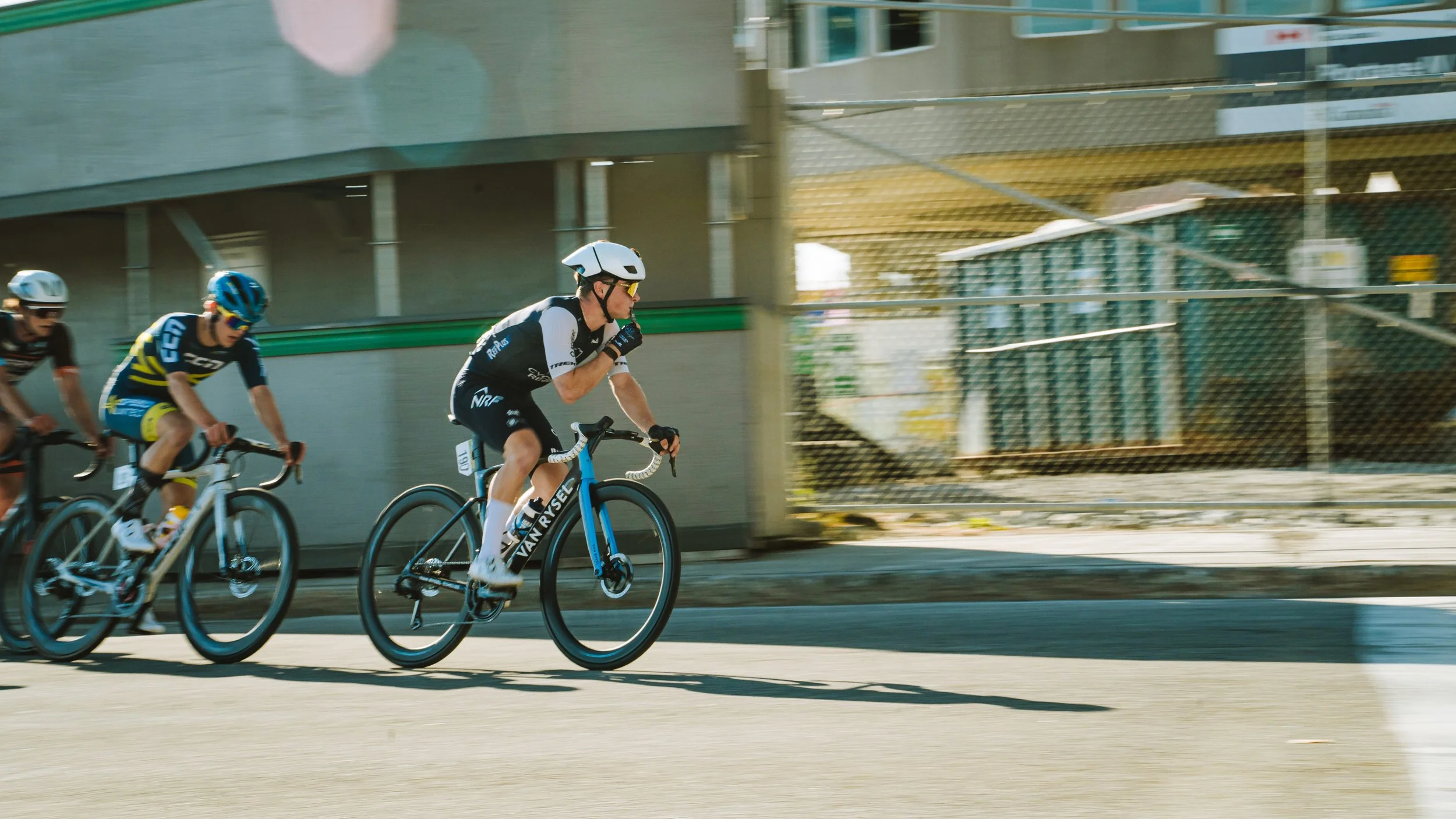 Three cyclists racing on a city street, wearing helmets and athletic gear, with a concrete wall and industrial storage in the background.
