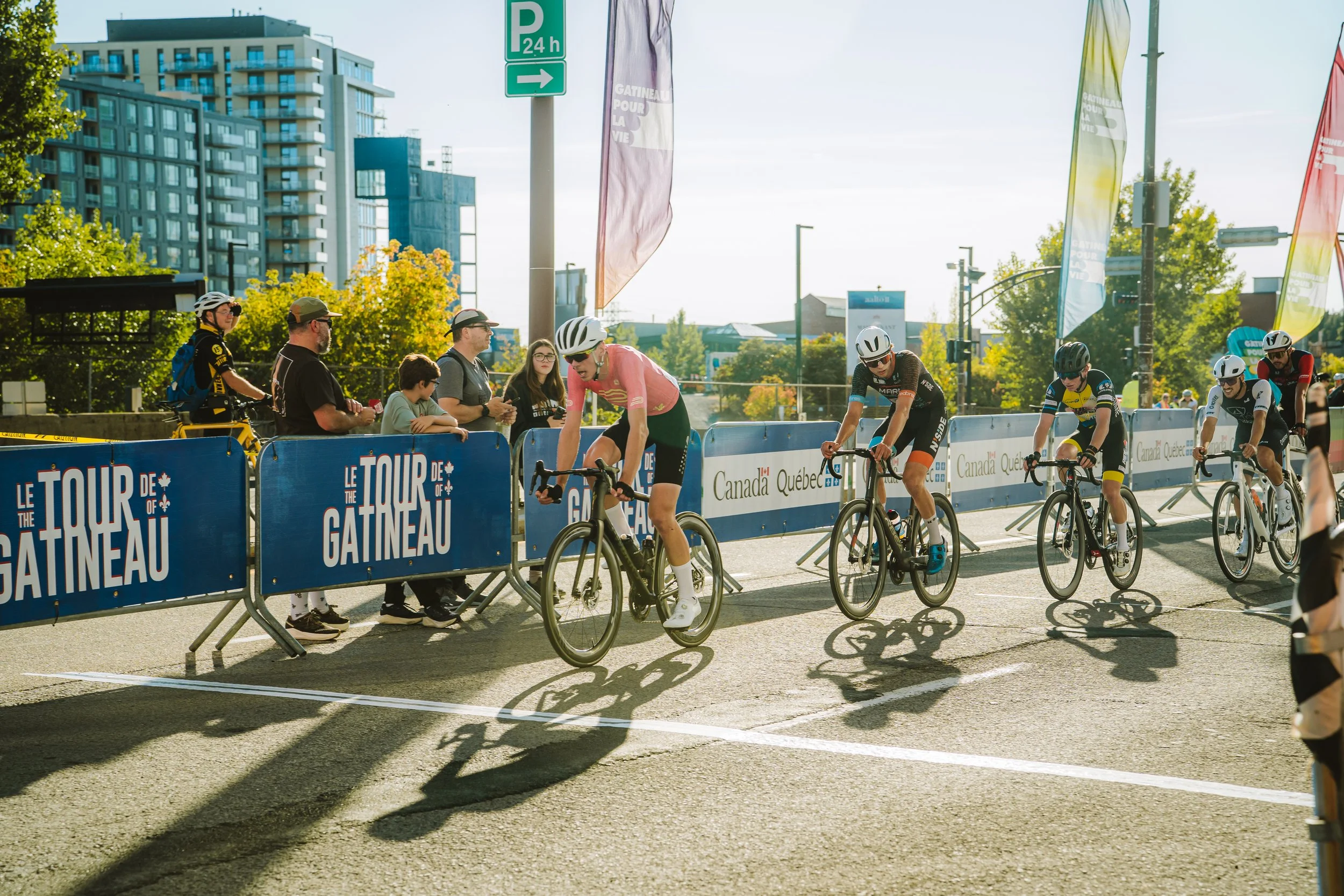 Cyclists crossing the finish line during a race at Le Tour de Gatineau, with spectators watching and urban buildings in the background.