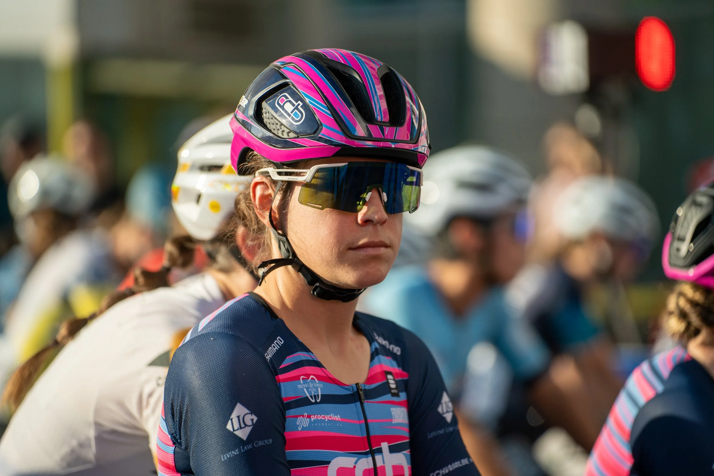A woman in cycling gear with a pink and blue helmet, wearing sunglasses, at a bike race or event.