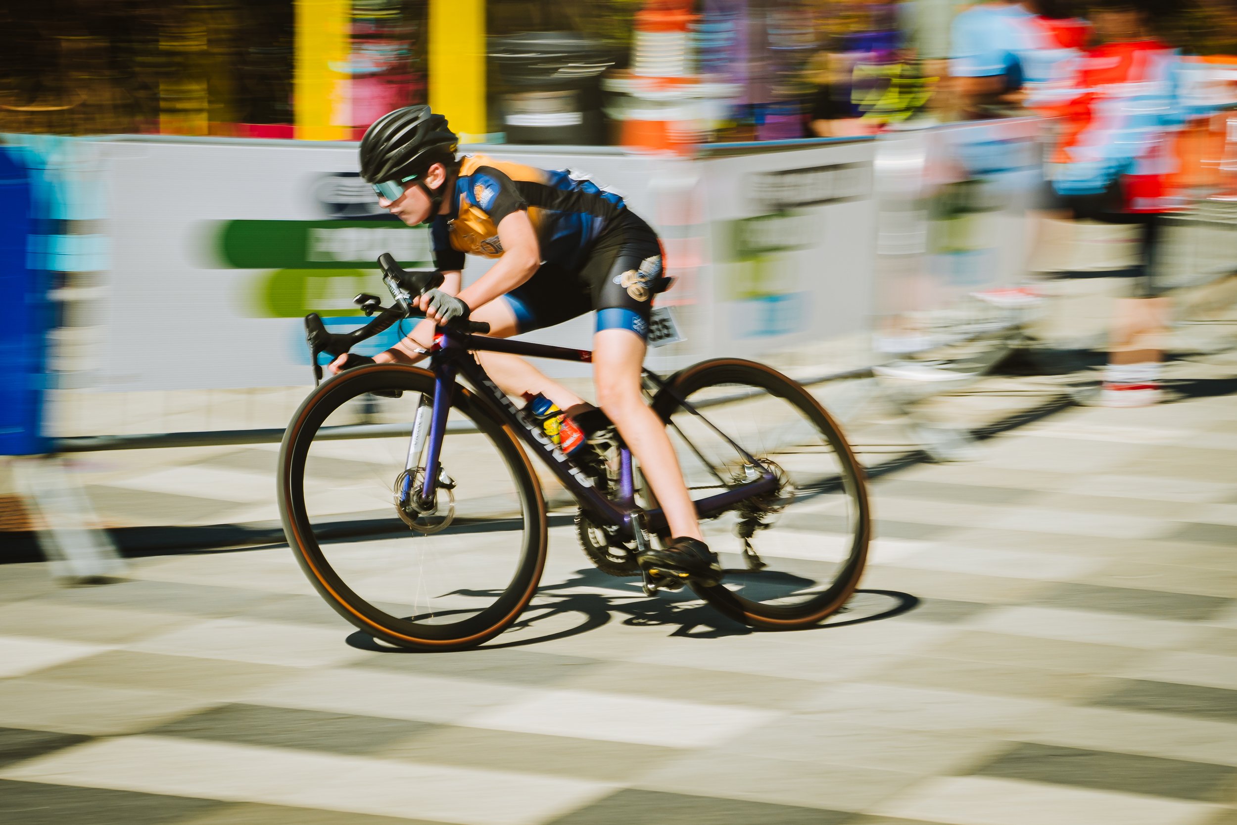 A cyclist in a black helmet and sports attire riding a black bicycle during a race, with blurred background and spectators.