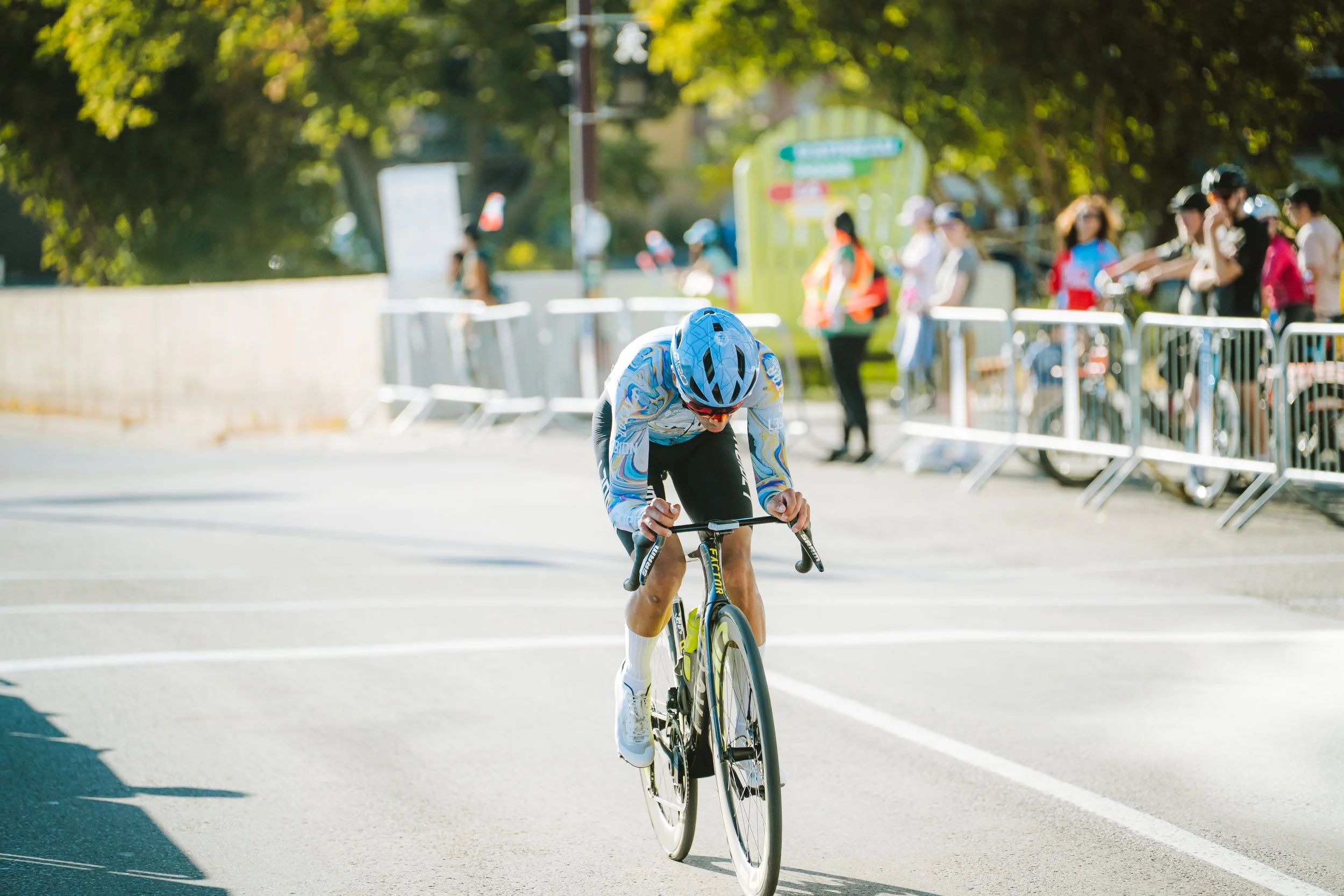 A cyclist in a blue helmet and colorful jersey riding a black and yellow road bike during a race, with spectators behind a metal barricade on a sunny day.