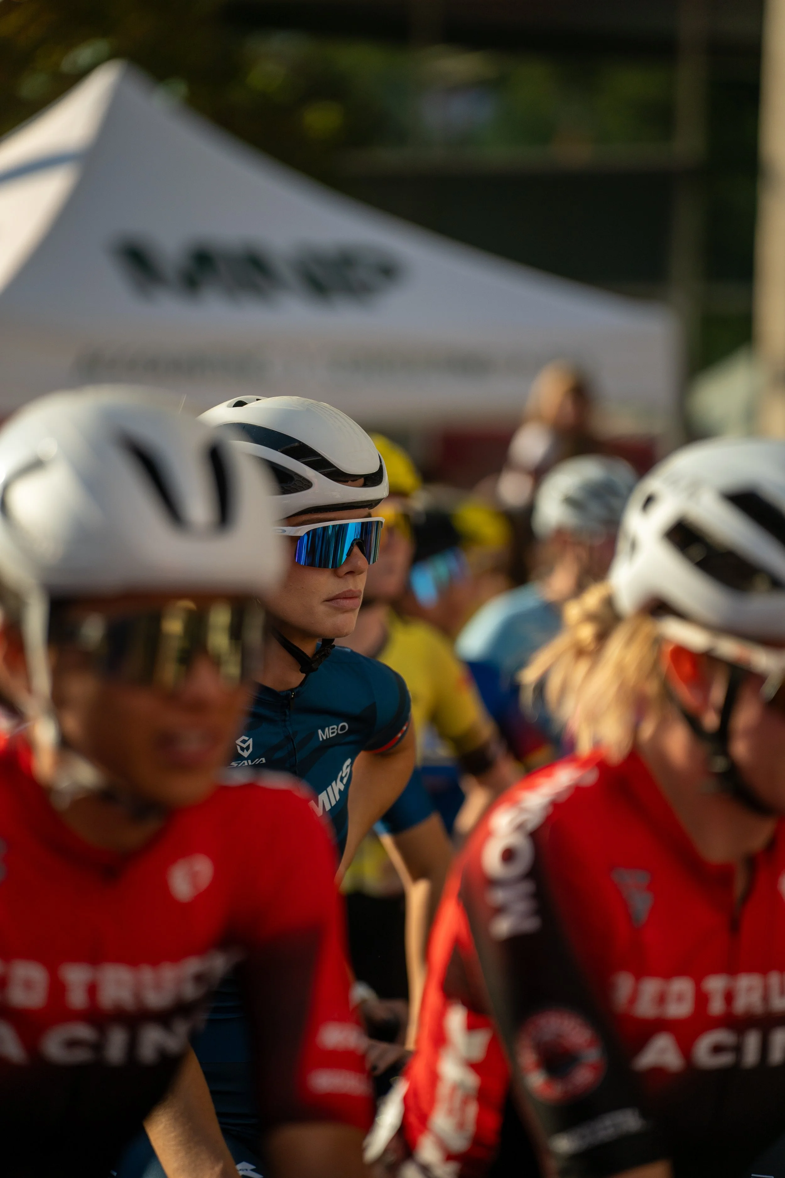 Multiple female cyclists wearing white helmets with black accents and sunglasses, standing in a row at a cycling event. There is a white tent with a blurred logo in the background.