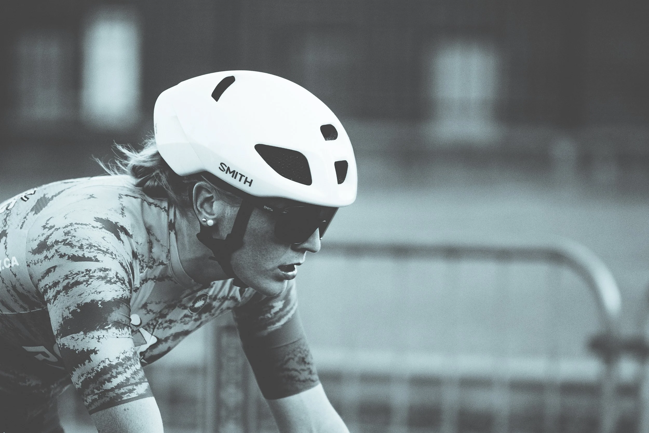 A woman wearing a white bike helmet, sunglasses, and a patterned cycling jersey, leaning forward as she rides a bicycle.