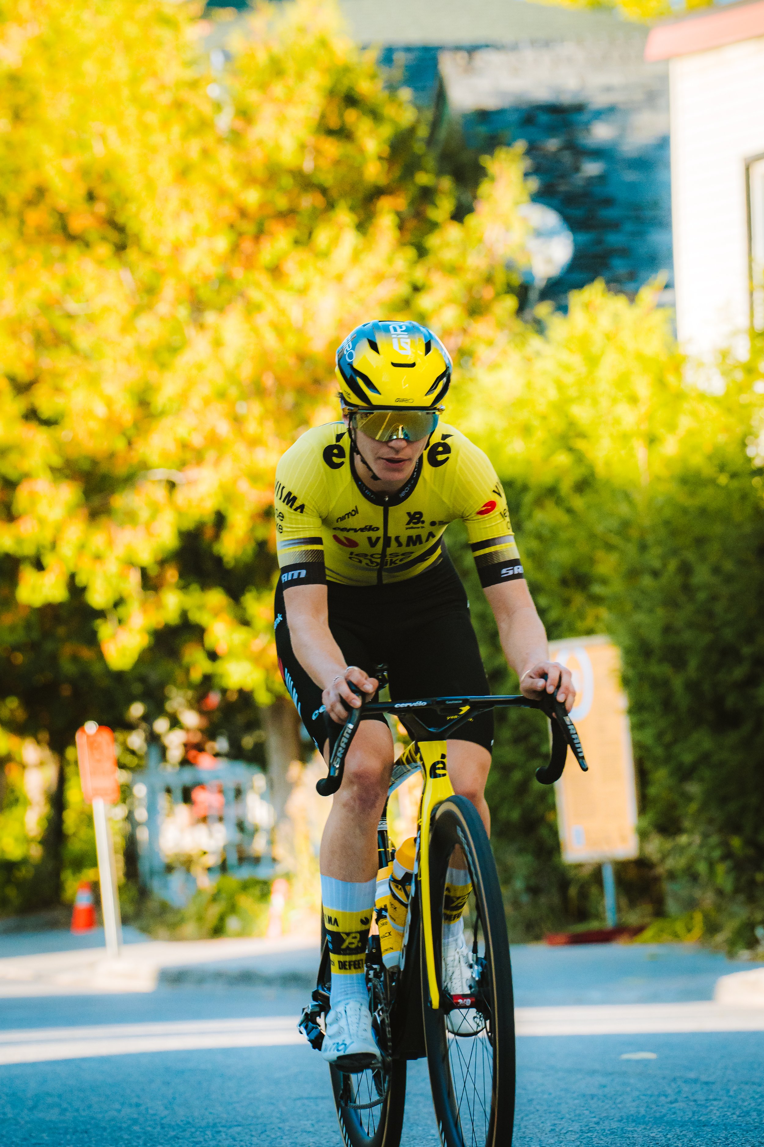 A cyclist in yellow and black riding a road bike on a street during daytime with trees and buildings in the background.