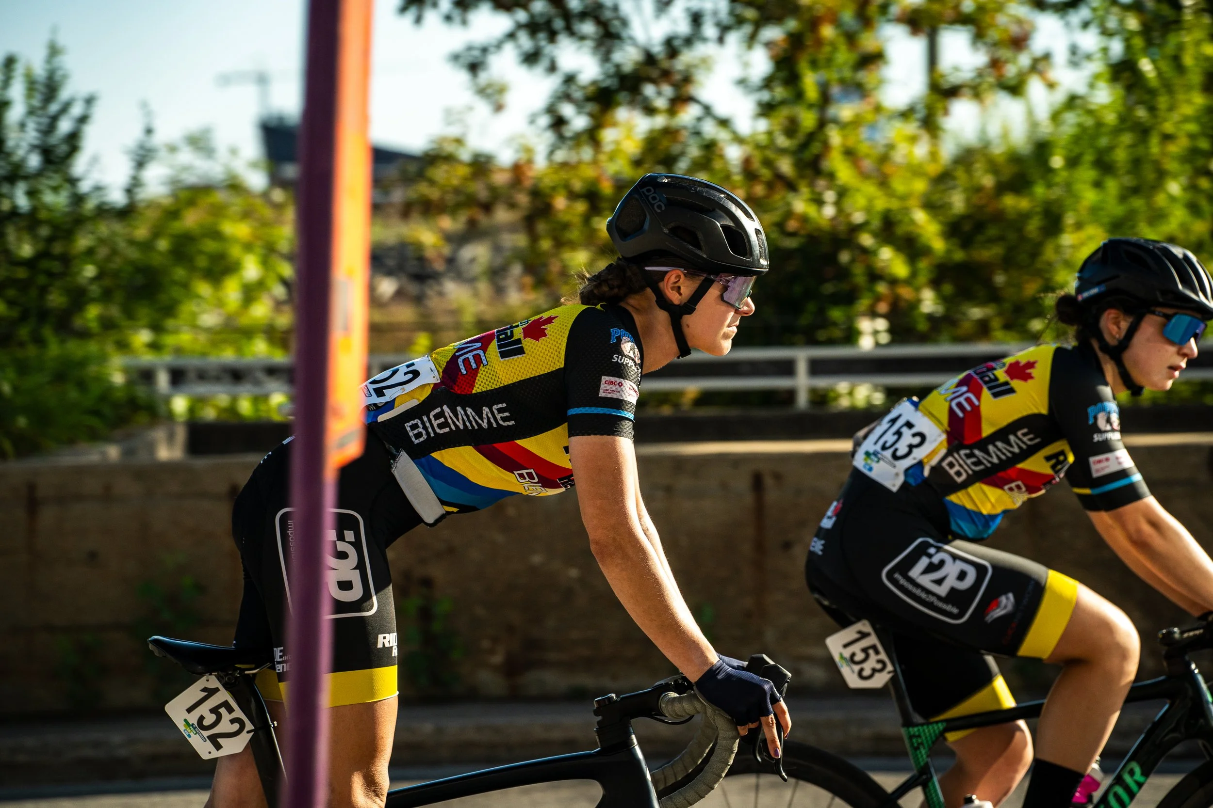 Two female cyclists wearing black helmets and colorful jerseys with the logo BIEMME, riding on a road during daytime with trees and a fence in the background.