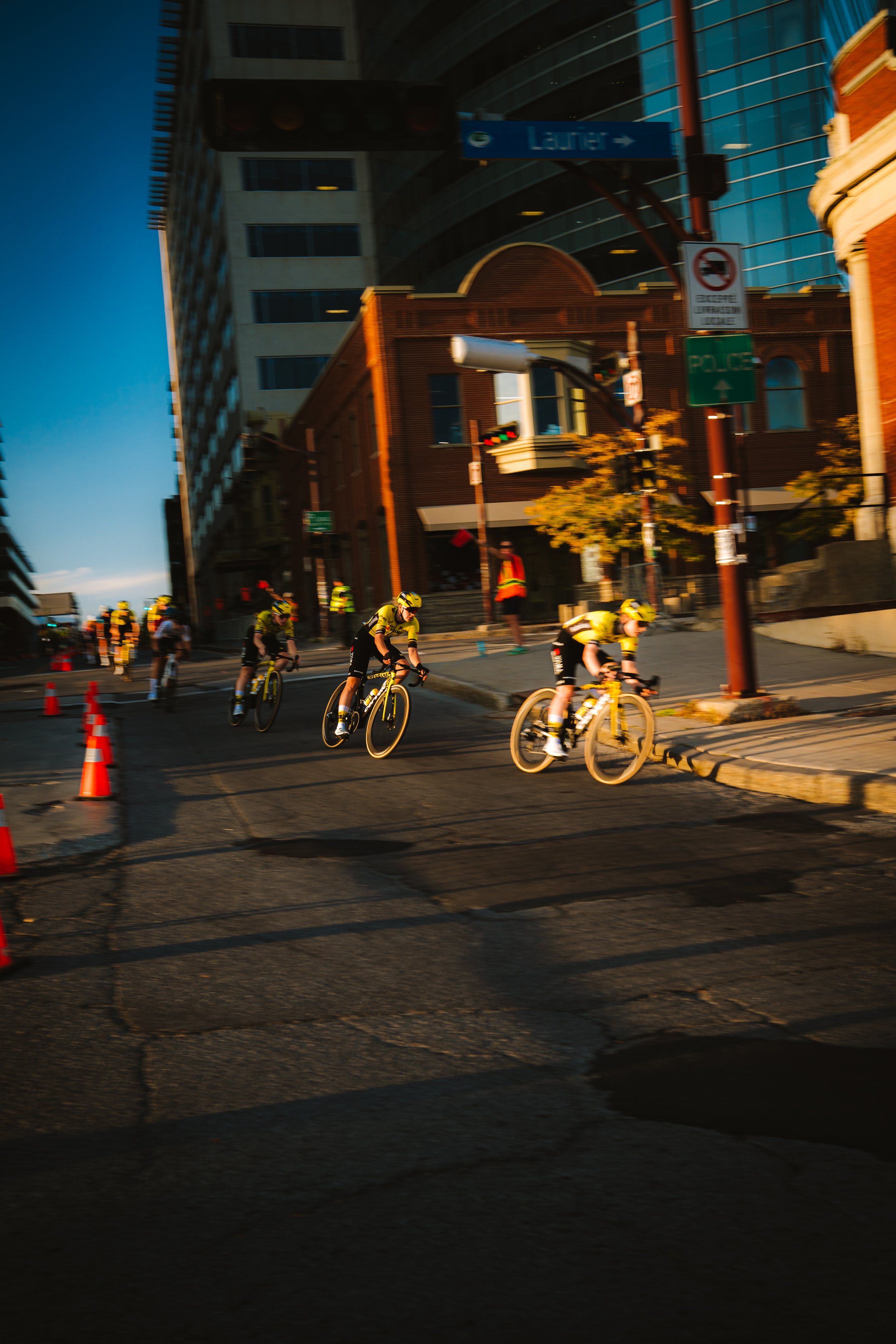 A group of cyclists riding downhill on a city street with orange traffic cones, surrounded by tall buildings and a pedestrian in a reflective vest, during late afternoon or early evening.