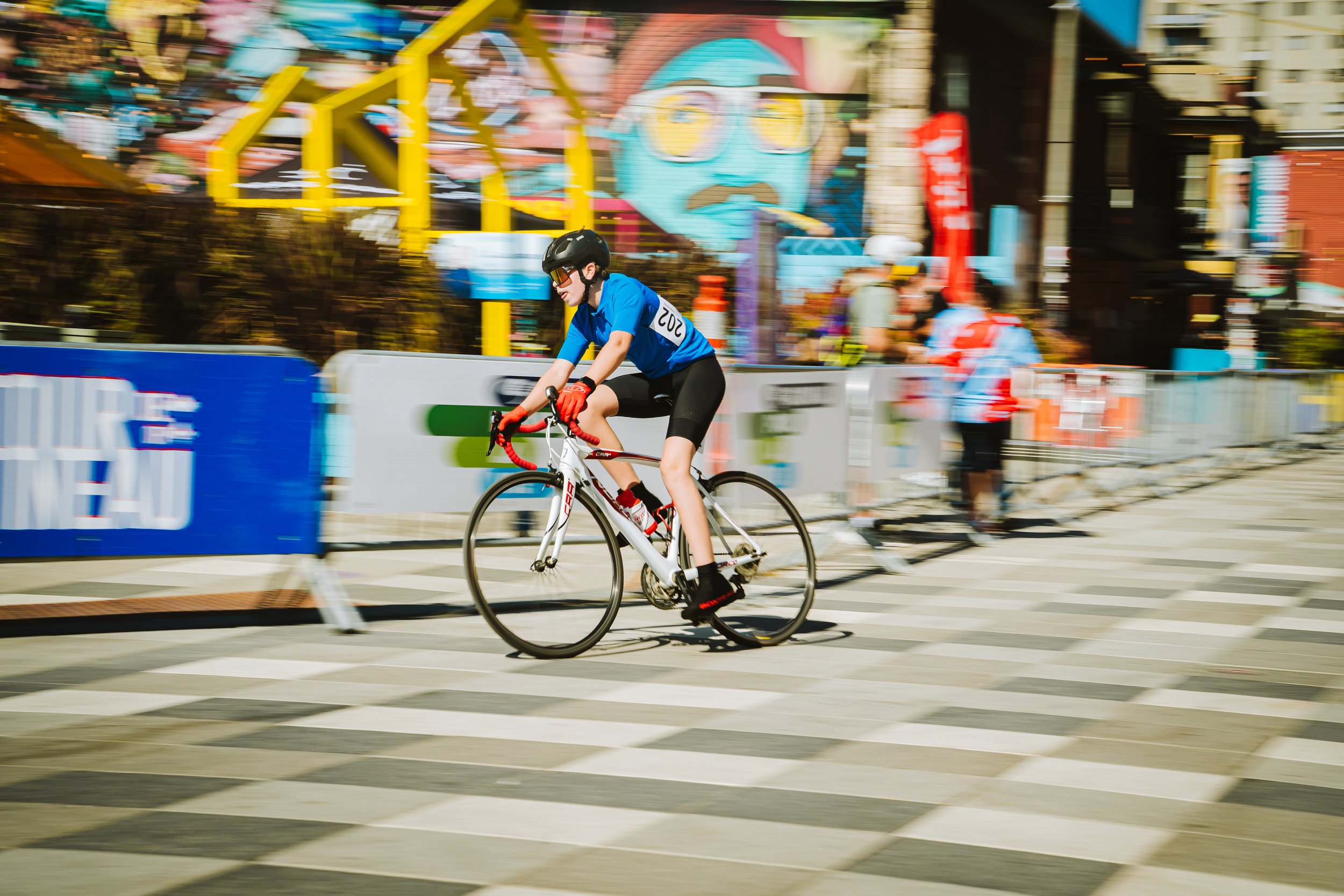 A cyclist riding a bike during a race or event in an urban area, with a colorful amusement park in the background.