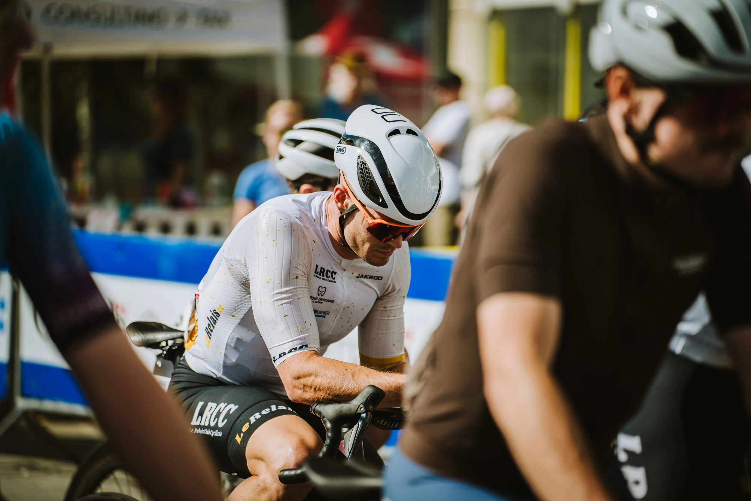 A cyclist in a white jersey and black shorts preparing for a race, wearing a white helmet and red sunglasses, surrounded by other cyclists and people at a race event.