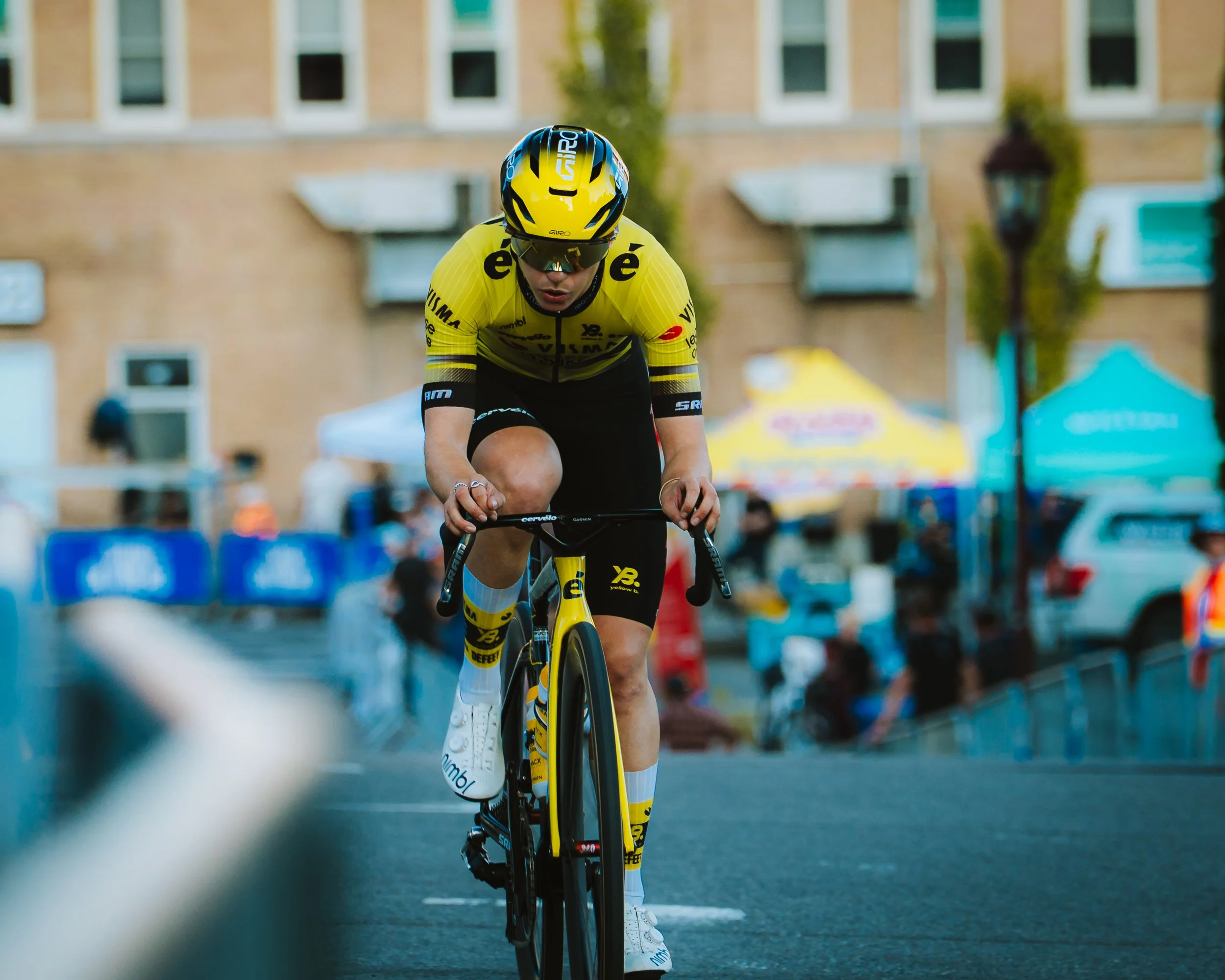 Cyclist in yellow and black uniform riding a racing bike on a street during a race, with tents and spectators in the background.