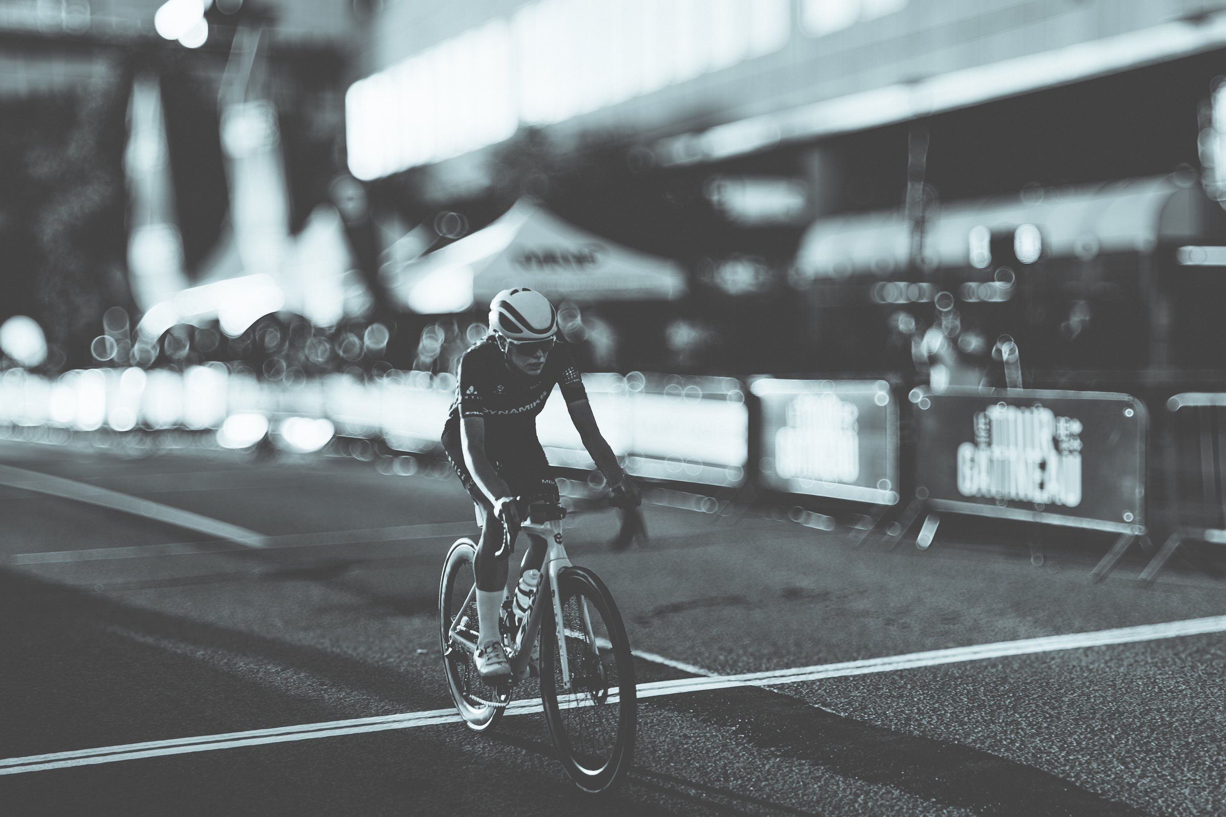 A cyclist riding a bike on a city street, wearing a helmet and sportswear, with barricades and blurred city lights in the background.