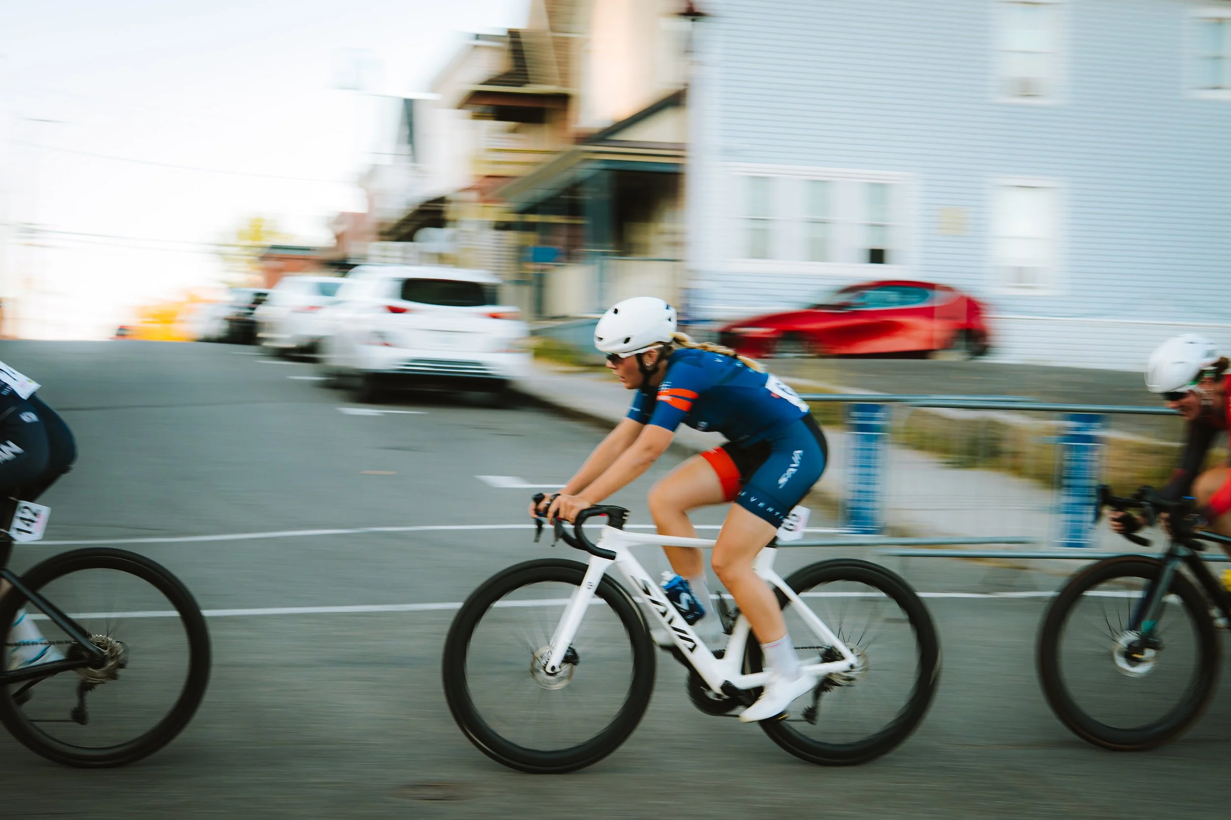 A woman cyclist participating in a race, riding a white bike wearing a white helmet, sunglasses, and a blue and red sports outfit, moving fast on a city street with blurred cars and buildings in the background.