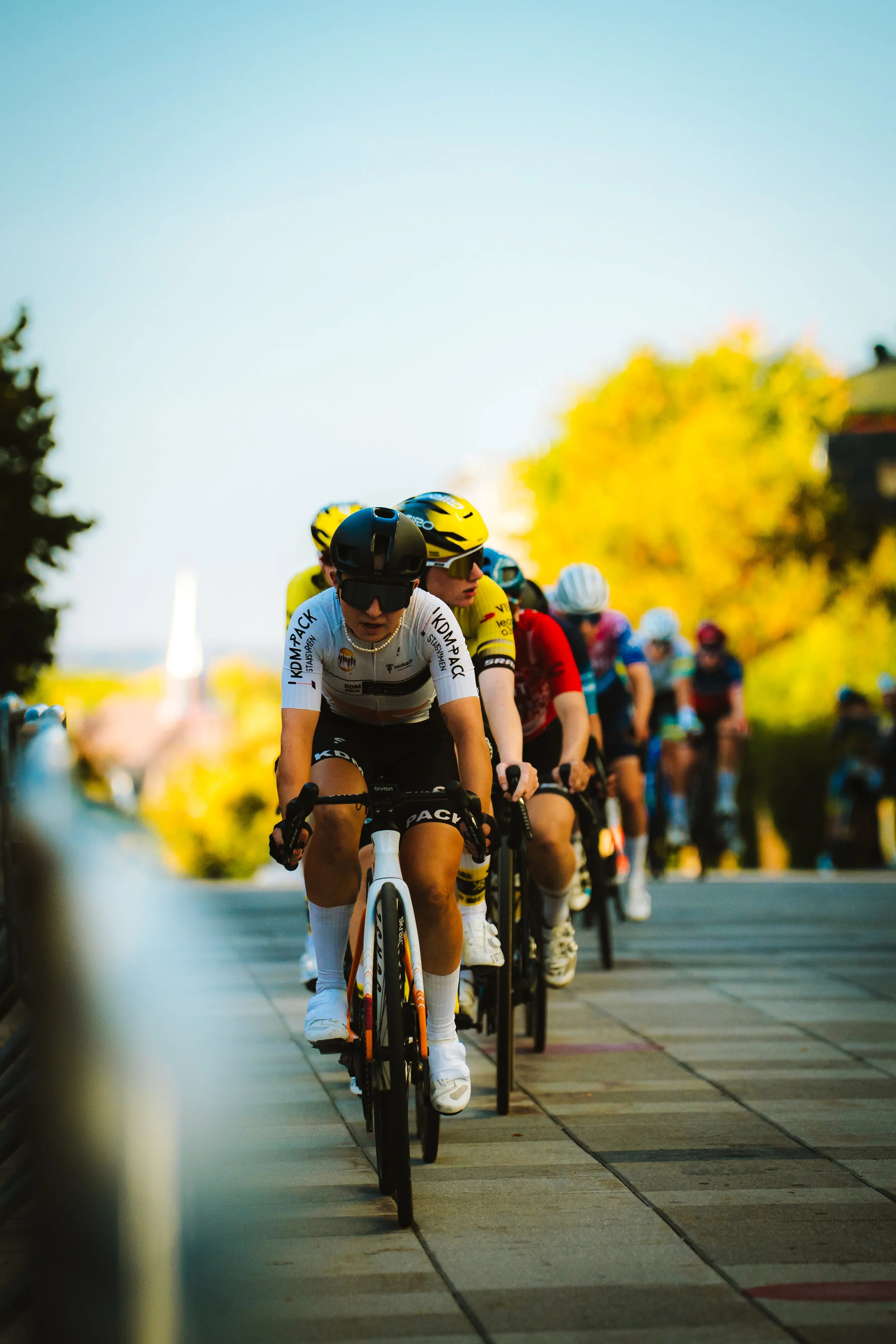Group of cyclists riding on a paved road during a race, with trees and colorful foliage in the background on a sunny day.