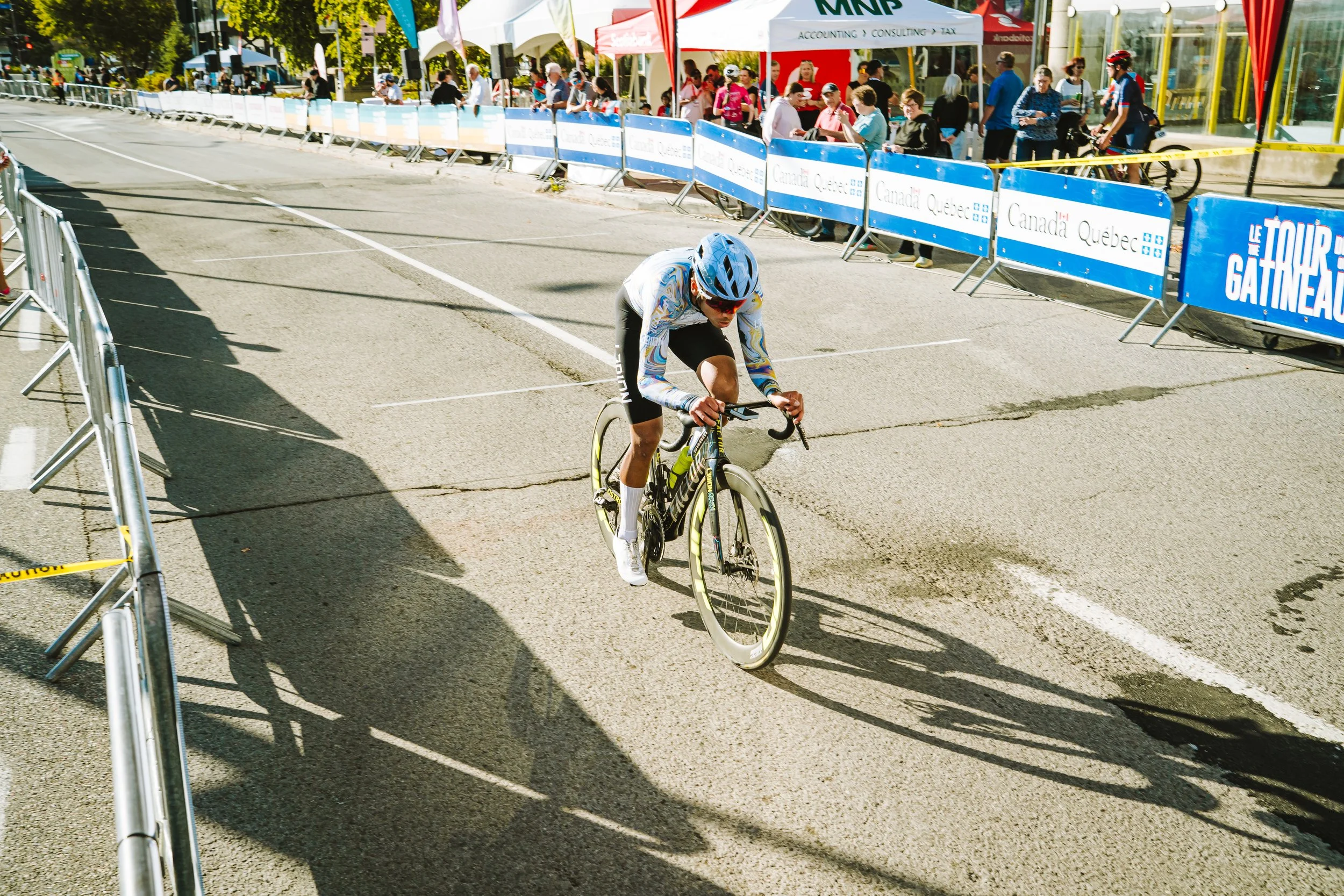 A cyclist racing on a street during a cycling event, with spectators behind safety barriers and a row of tents in the background.