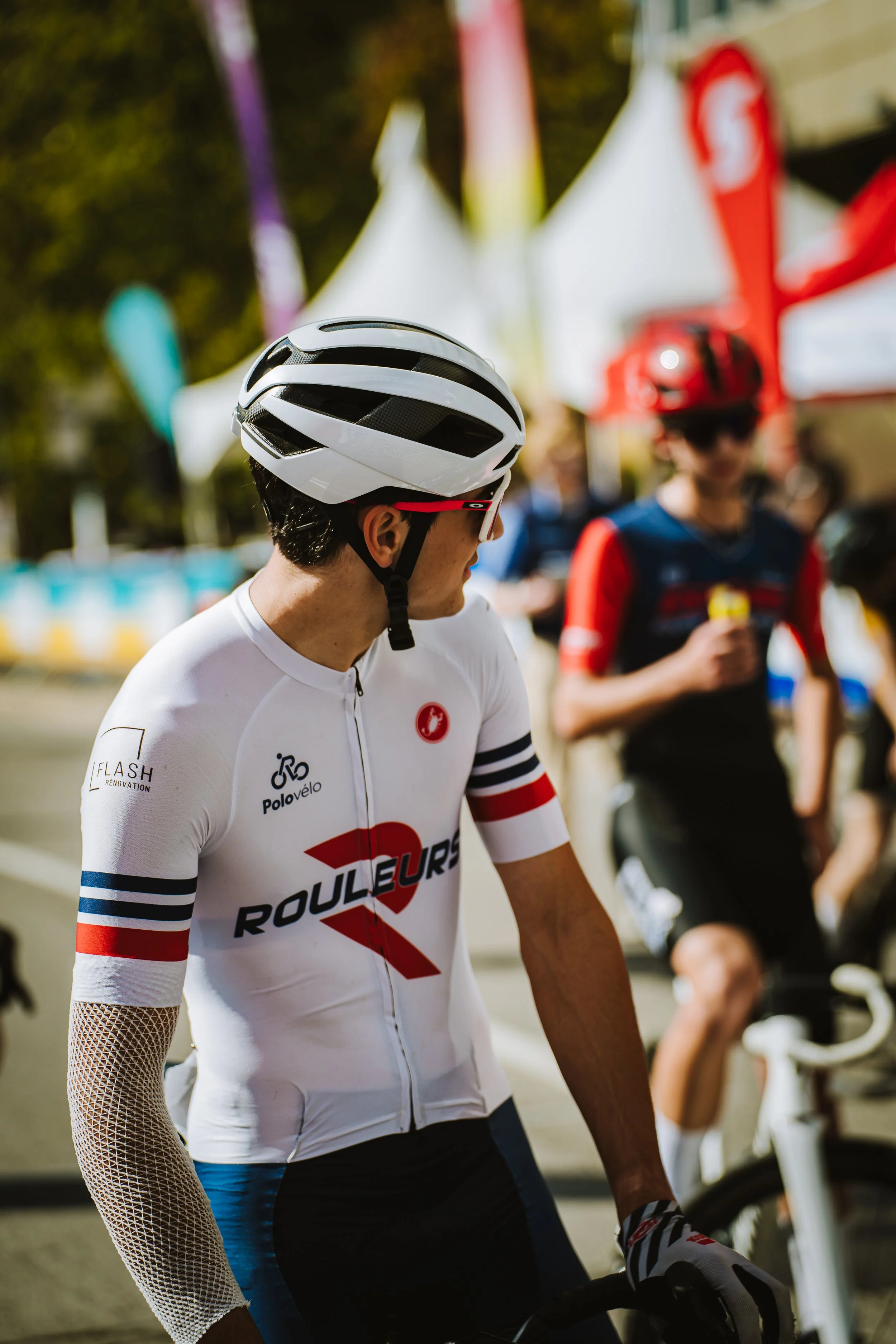Cyclist wearing a white helmet and a white jersey with red and blue accents, standing with a bicycle at a cycling event.
