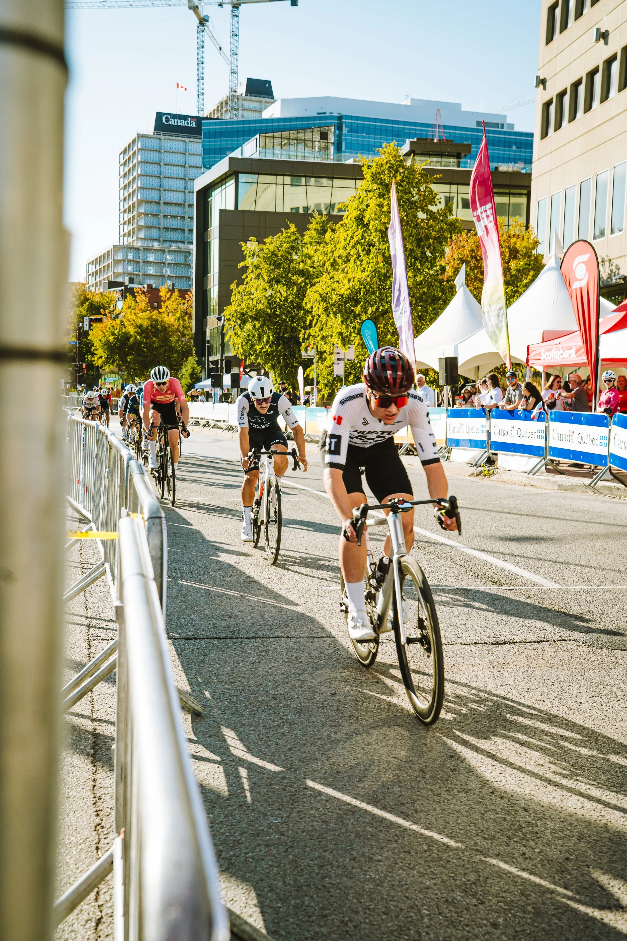 Cyclists race on a city street in a competitive cycling event, with spectators and flags along the sidewalk, and modern buildings in the background.