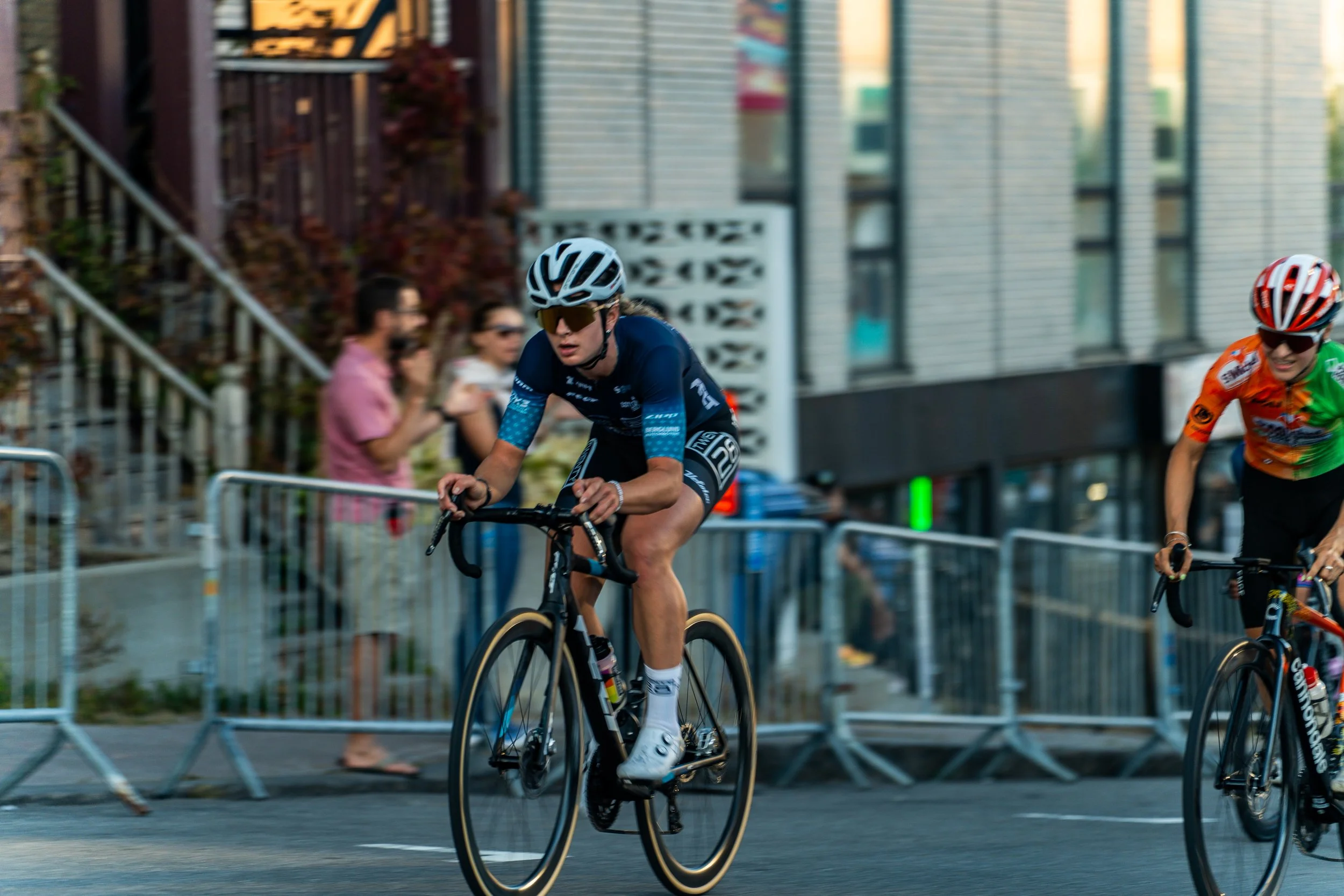 Cyclists in a race, wearing helmets and athletic gear, riding on city streets with spectators and barriers in the background.