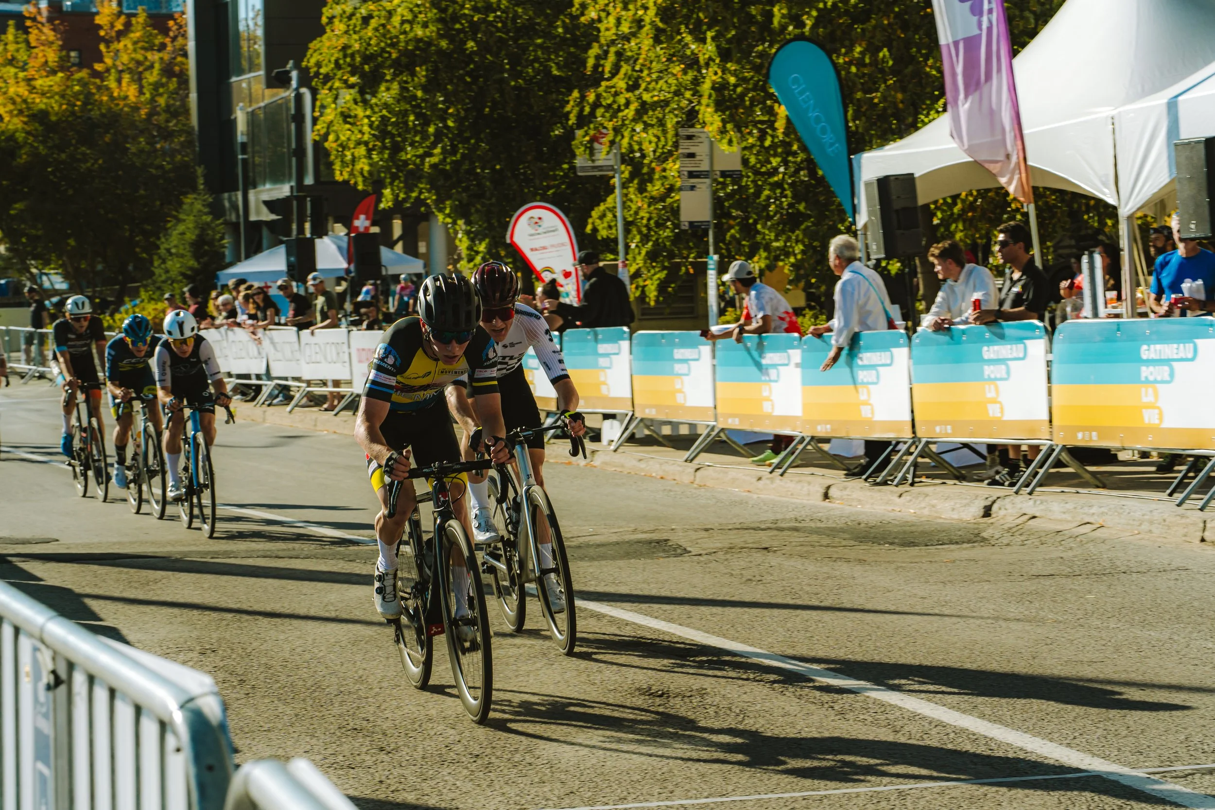 Cyclists racing on a city street during a competition with spectators and roadside banners.