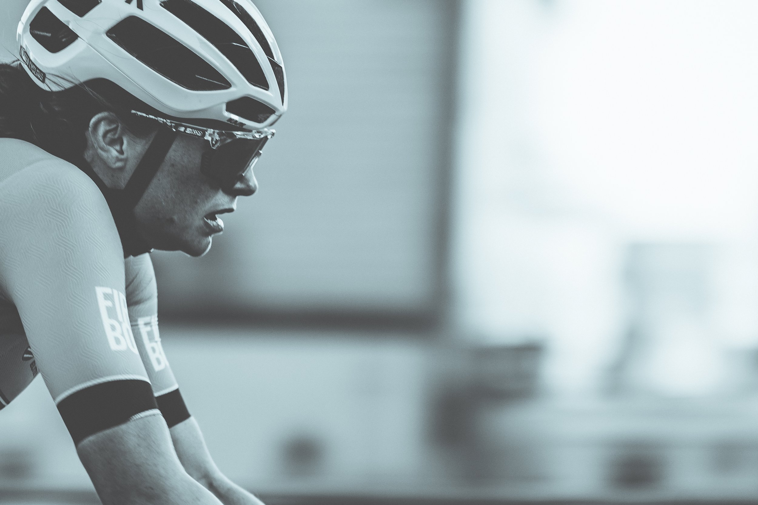 A cyclist wearing a helmet, sunglasses, and athletic gear, leaning forward with a focused expression, in a monochrome setting.