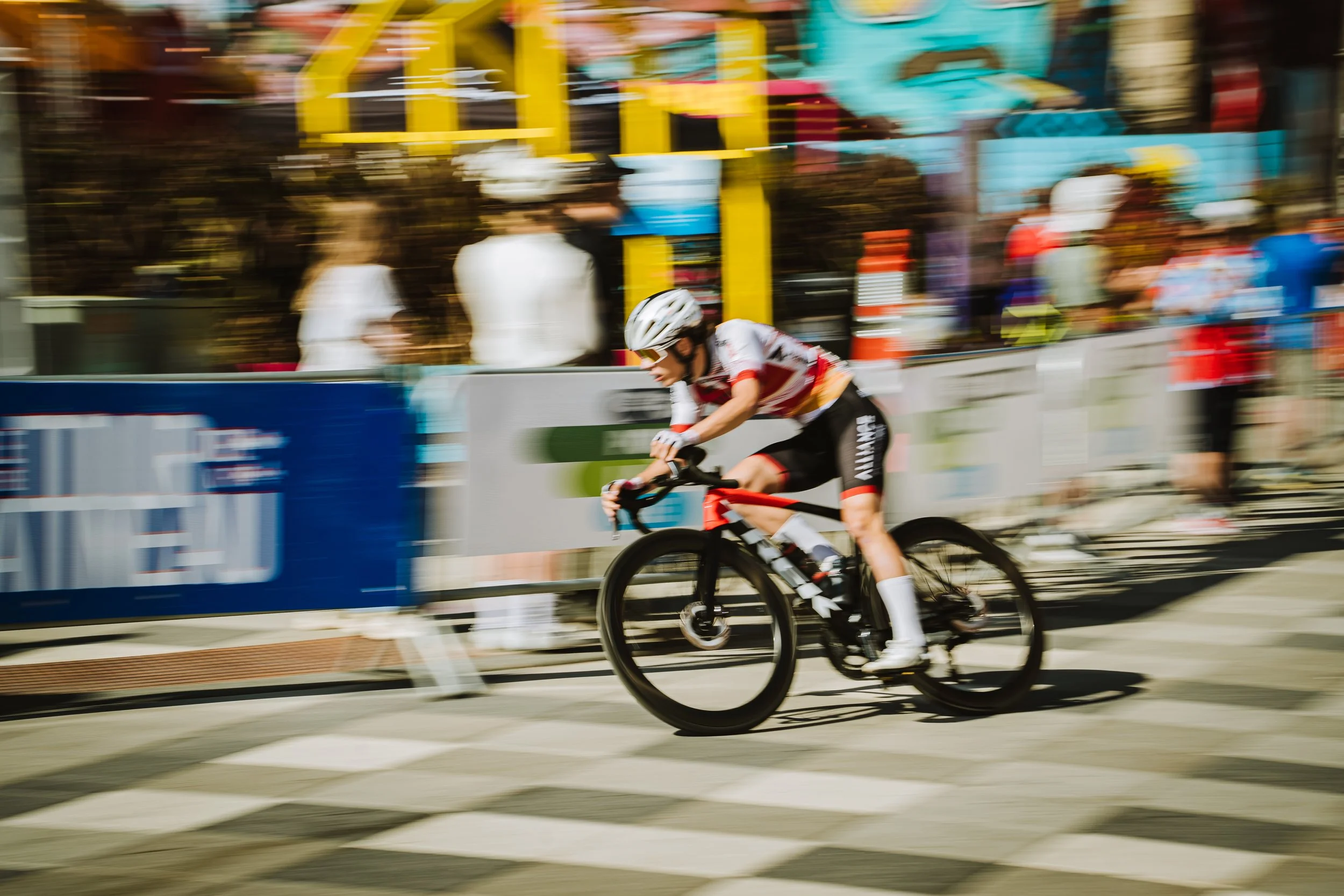 A cyclist wearing a white helmet and sports gear riding a black and red bicycle during a race, with blurred spectators and colorful structures in the background.