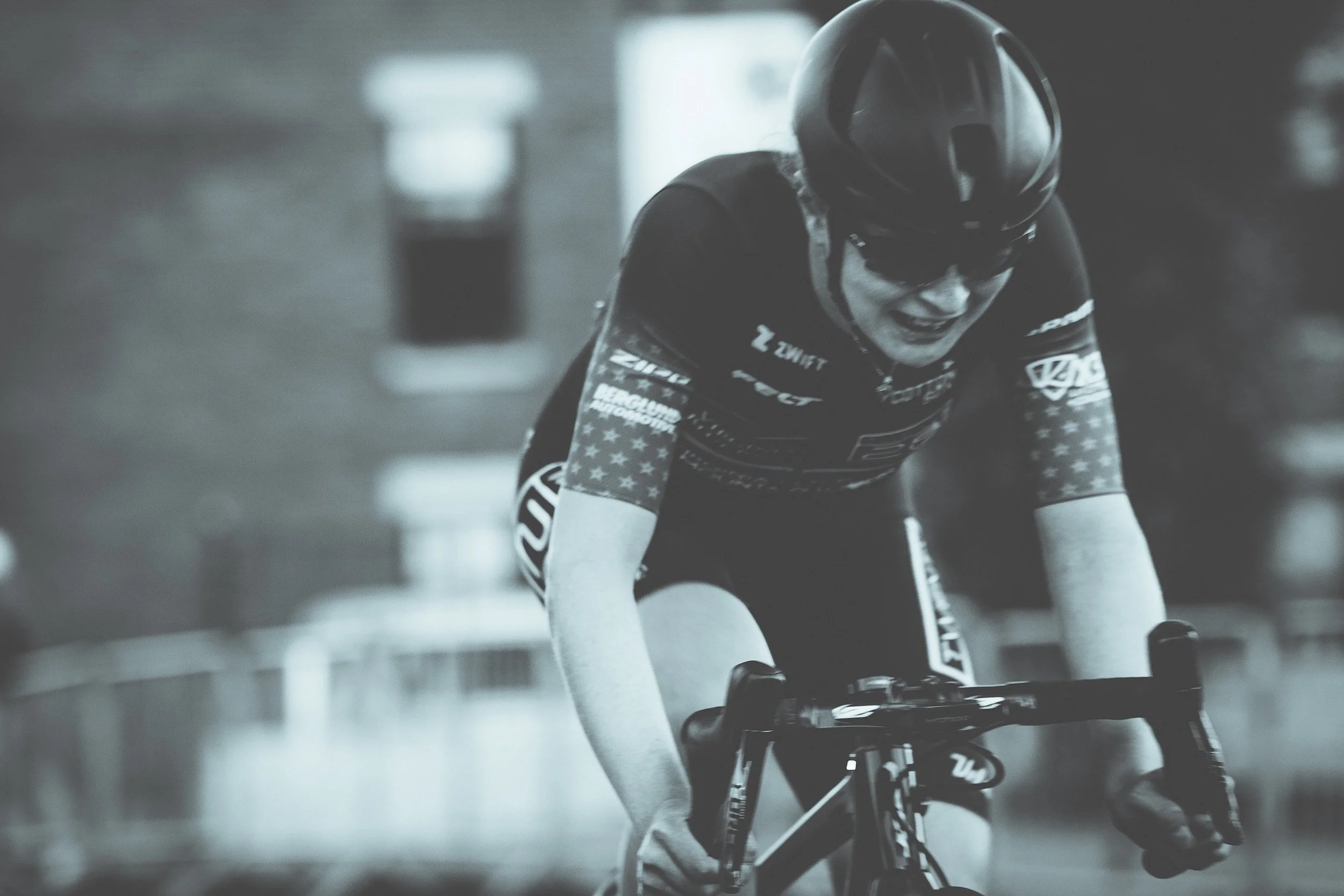 A cyclist in a black helmet and cycling outfit leaning forward while riding a bike indoors, with a focused expression.