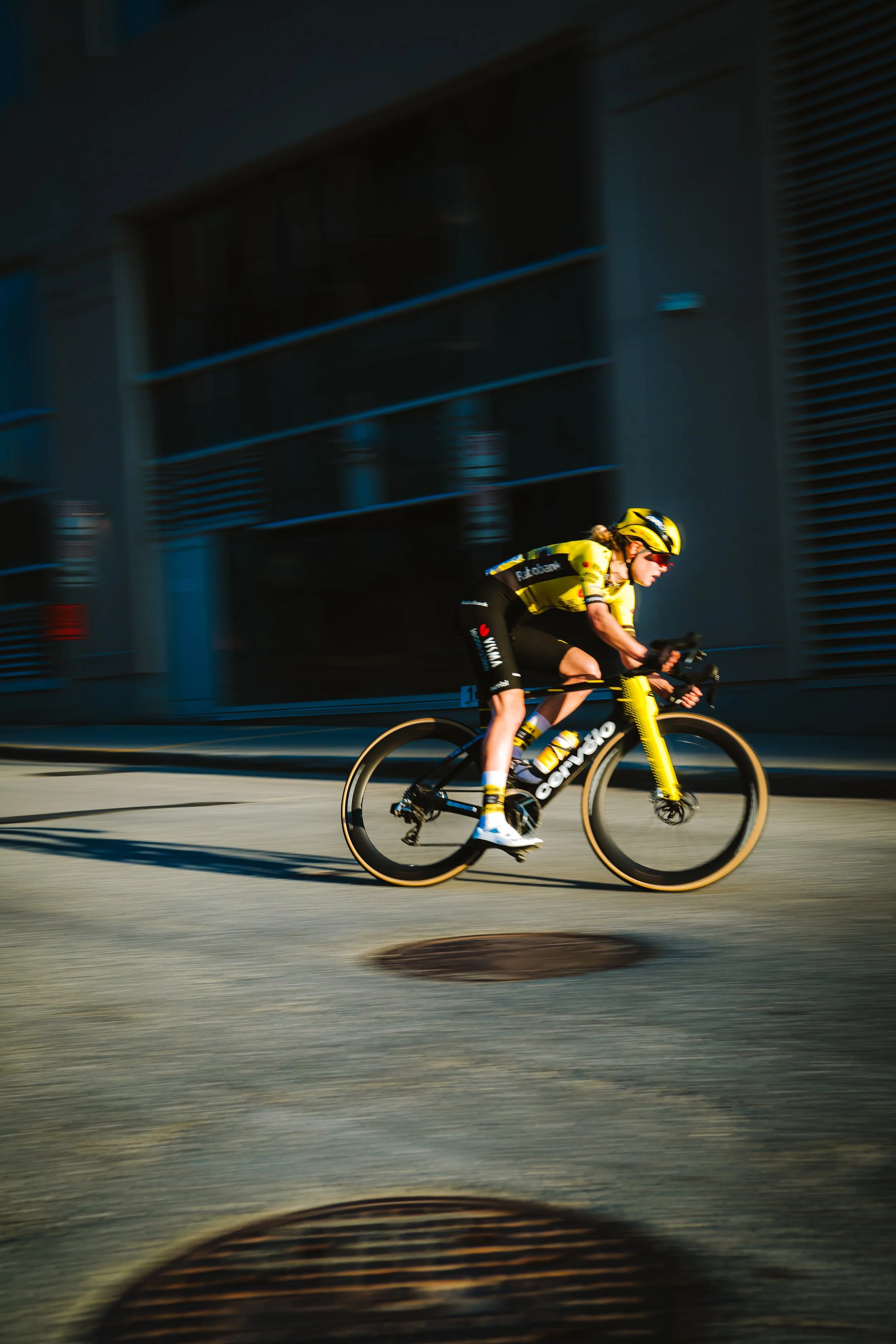 A cyclist in yellow and black gear riding a black and yellow racing bike at high speed on an urban street during daytime.