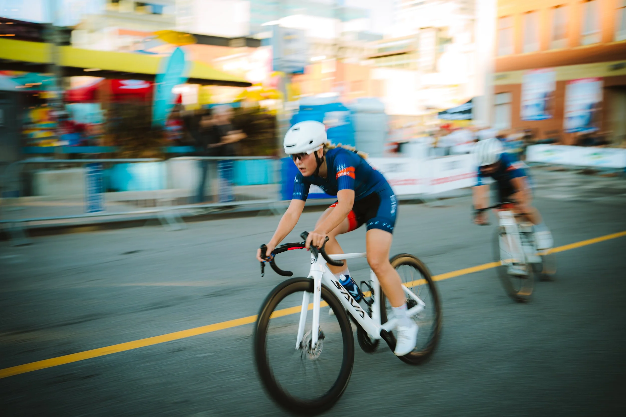 A woman wearing a white helmet and blue cycling outfit riding a white road bike during a race with a blurred background of colorful buildings and another cyclist.