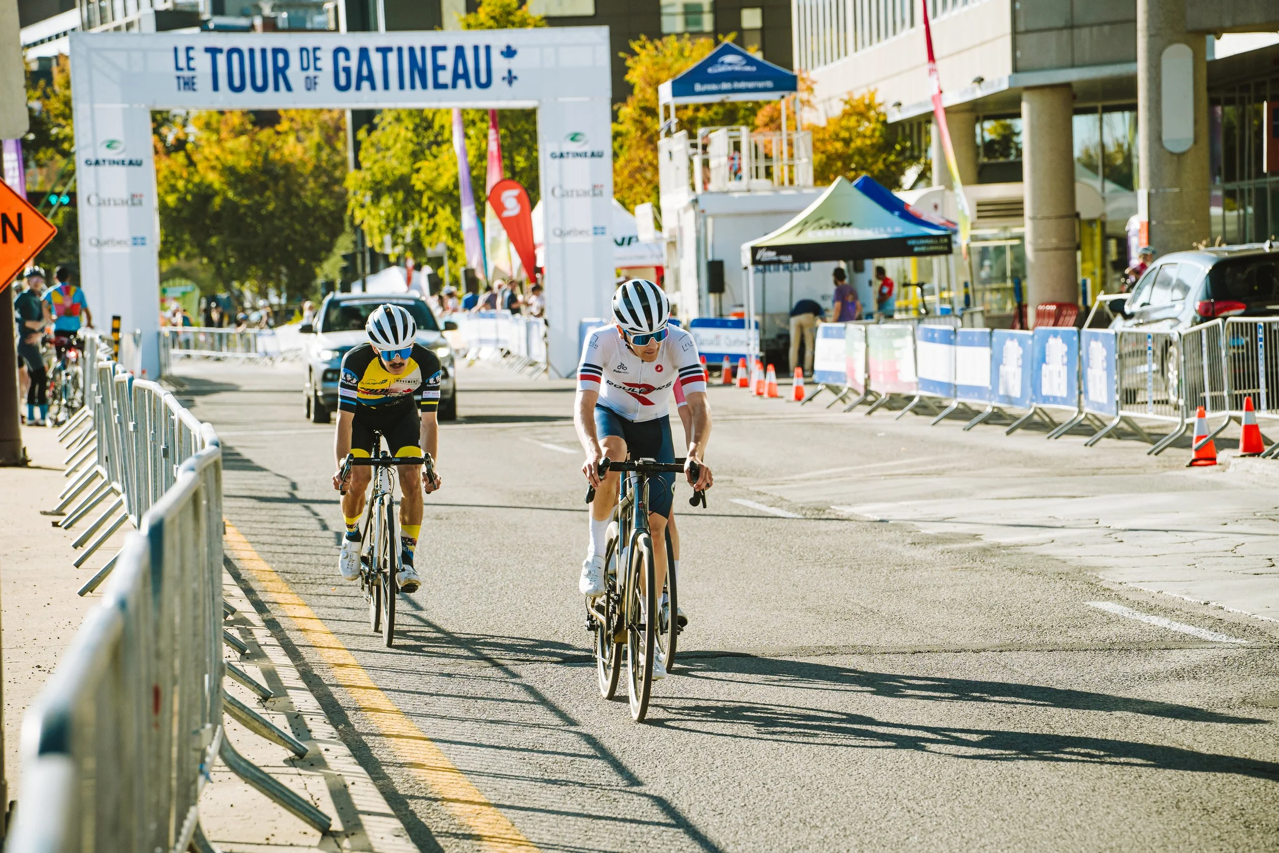 Two cyclists racing in a city street during a cycling event, with a large banner overhead that reads 'Le Tour de Gatineau' and barriers lining the road.