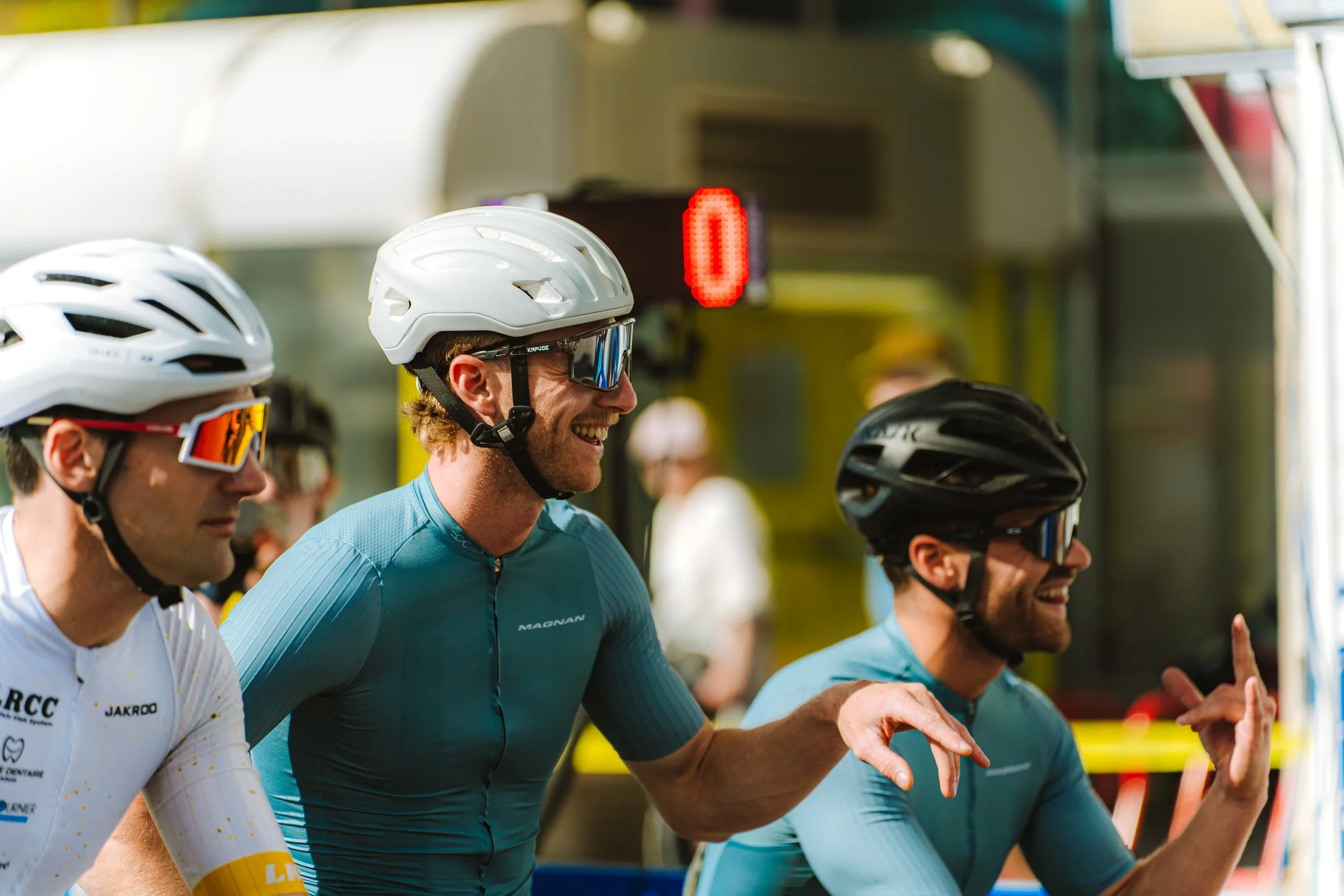 Three cyclists in racing gear and helmets smiling and interacting at a race event.