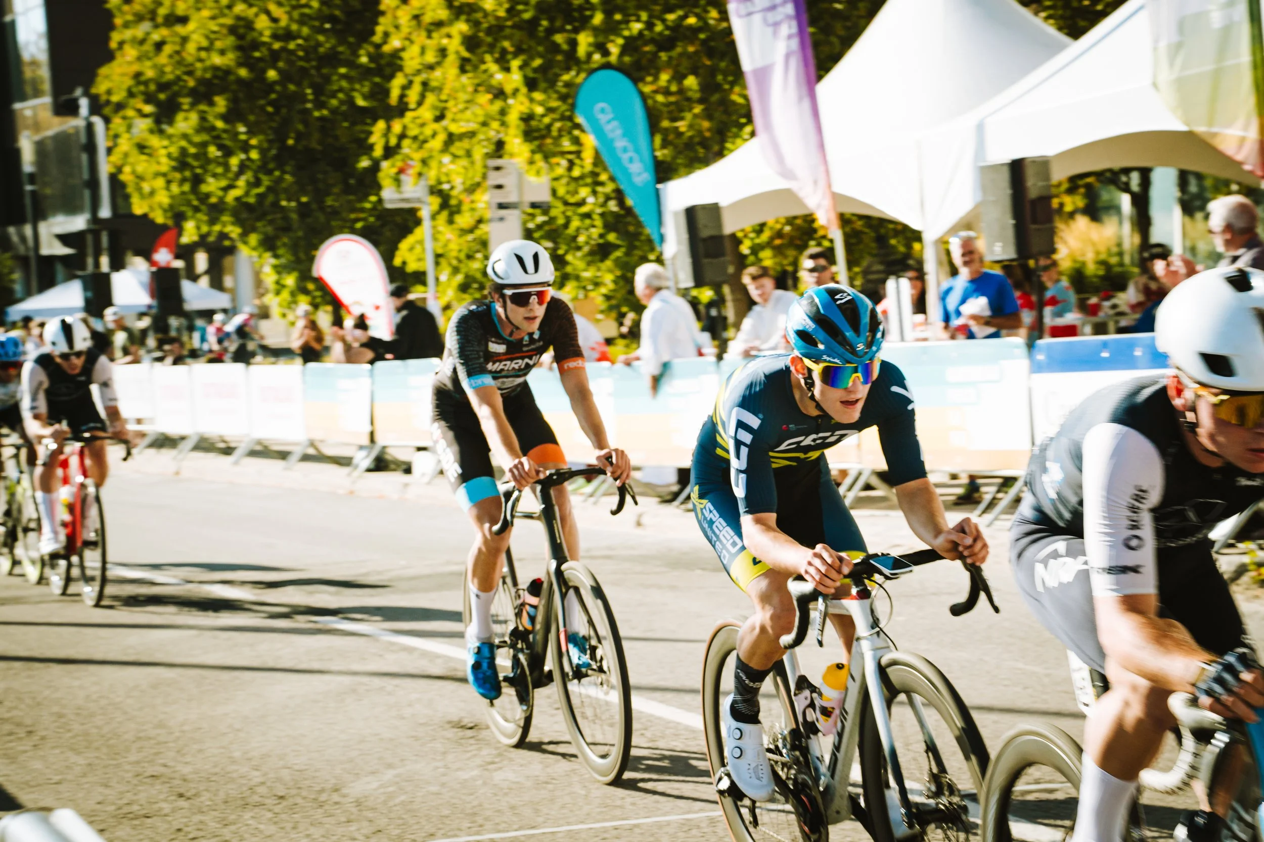 Cyclists competing in a race on a city street, with spectators and banners in the background.
