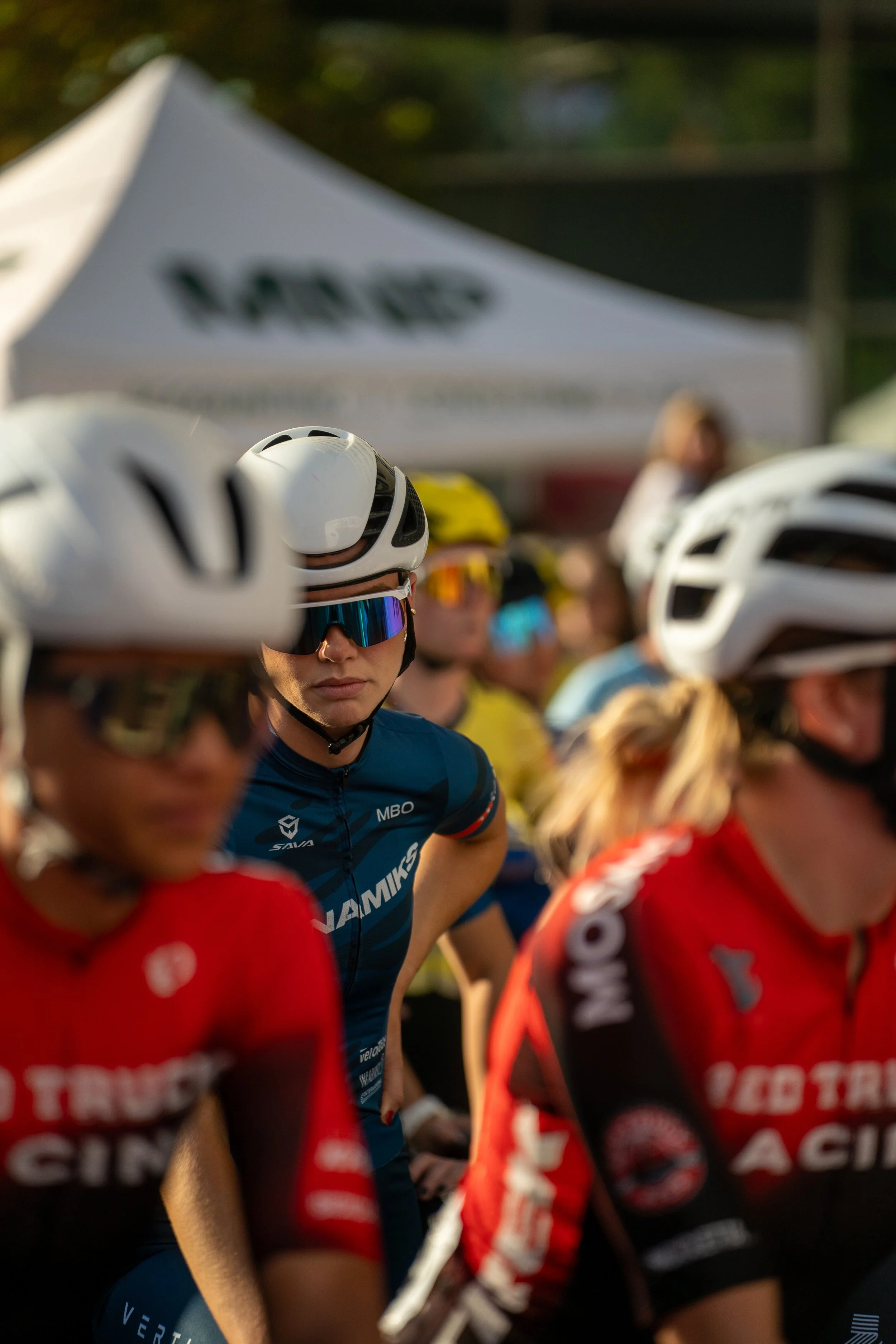 Cyclists wearing helmets and sunglasses at the start line of a race, with a white tent in the background.