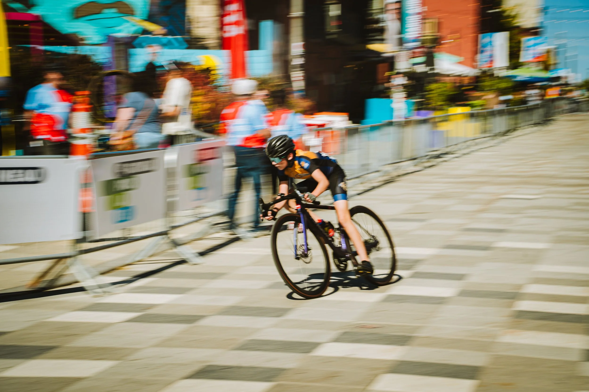 A young cyclist riding a bicycle on a city street during a race or event, with a blurred background of pedestrians, colorful buildings, and barriers.