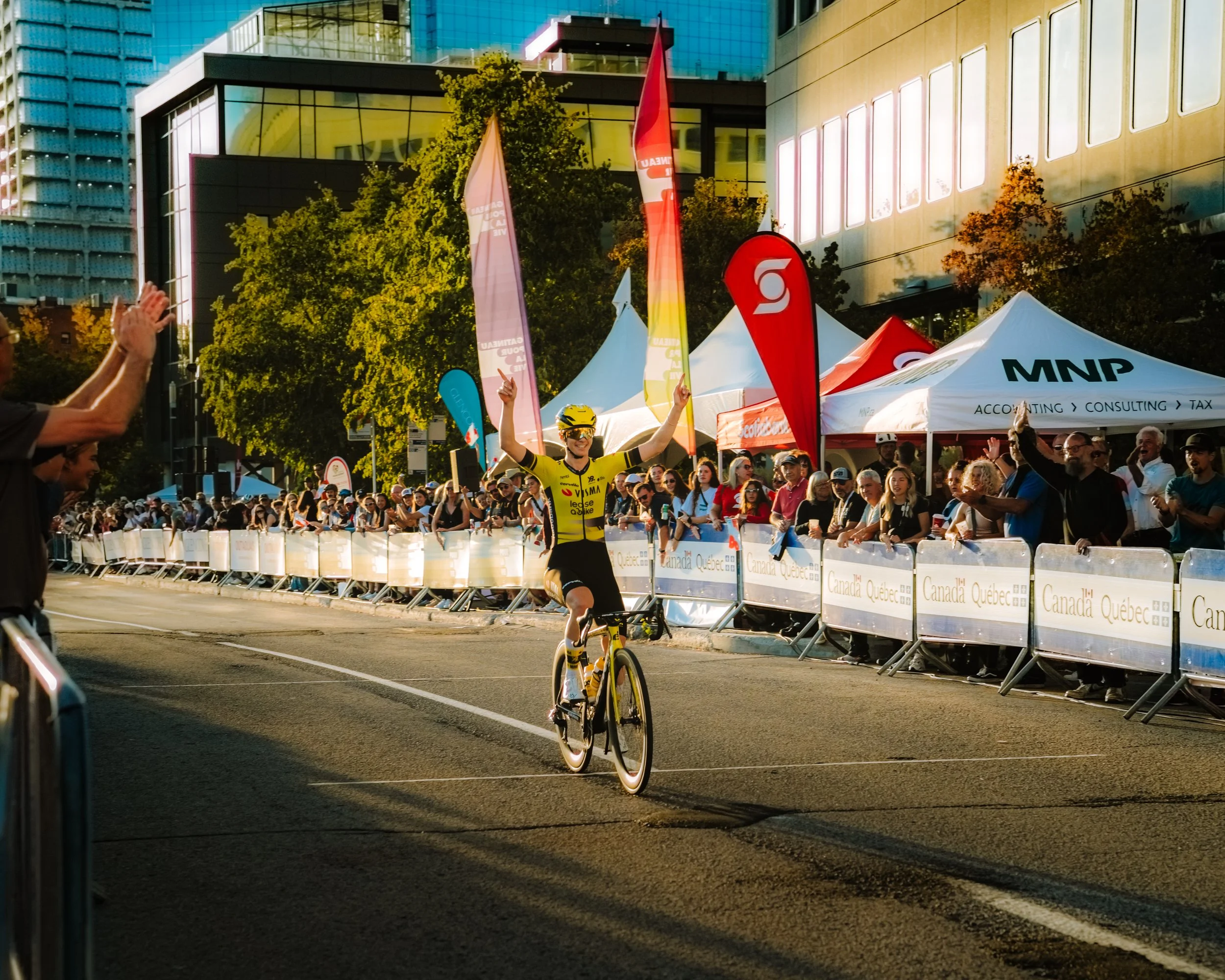 A cyclist in a yellow team jersey celebrating as he crosses the finish line during a race, surrounded by cheering spectators behind barricades, with tents and flags in the background.