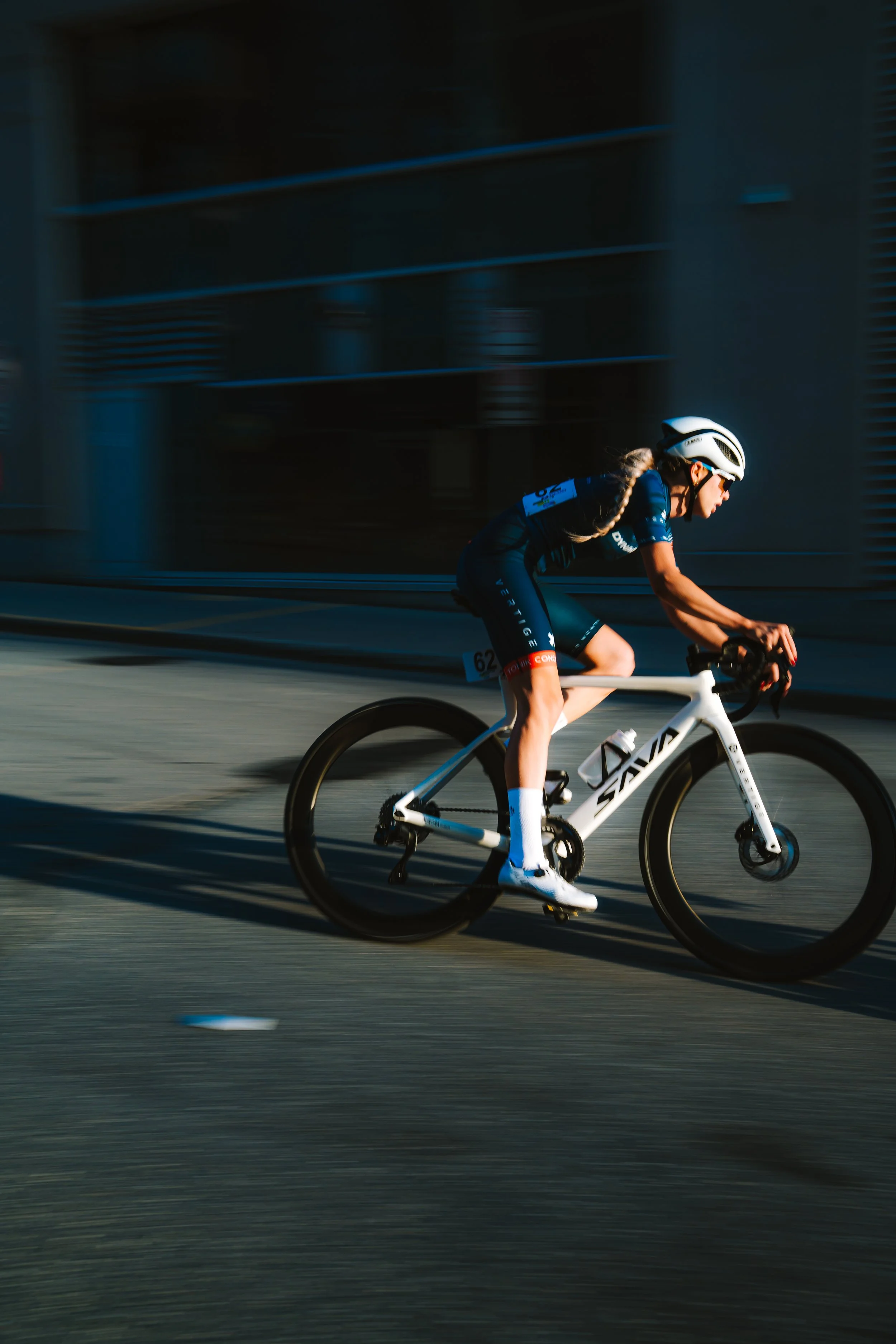 A female cyclist wearing a helmet, sunglasses, and a dark cycling outfit riding a white road bike on a city street during daytime.