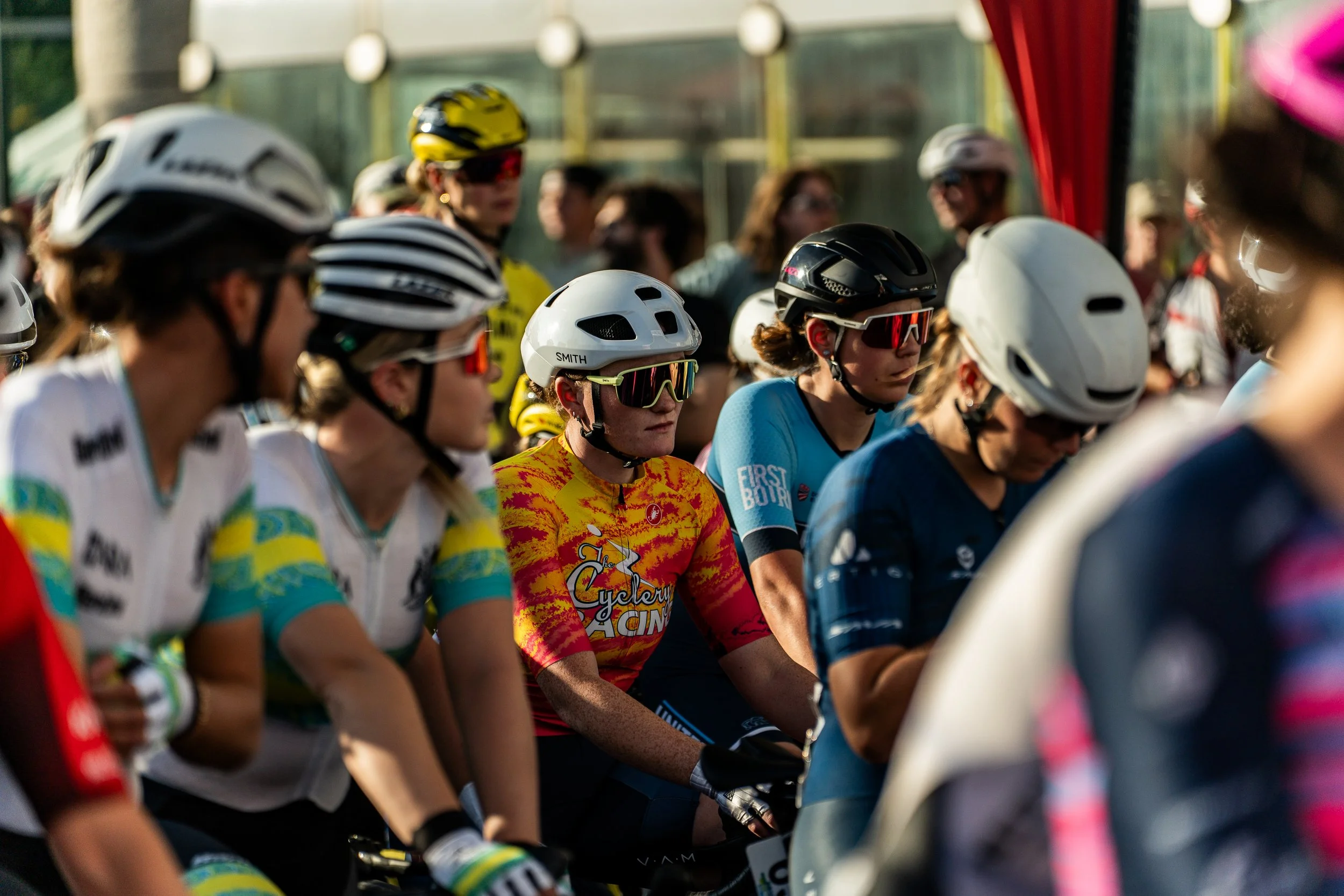 Group of female cyclists at the starting line, wearing helmets and colorful jerseys, ready for a race.