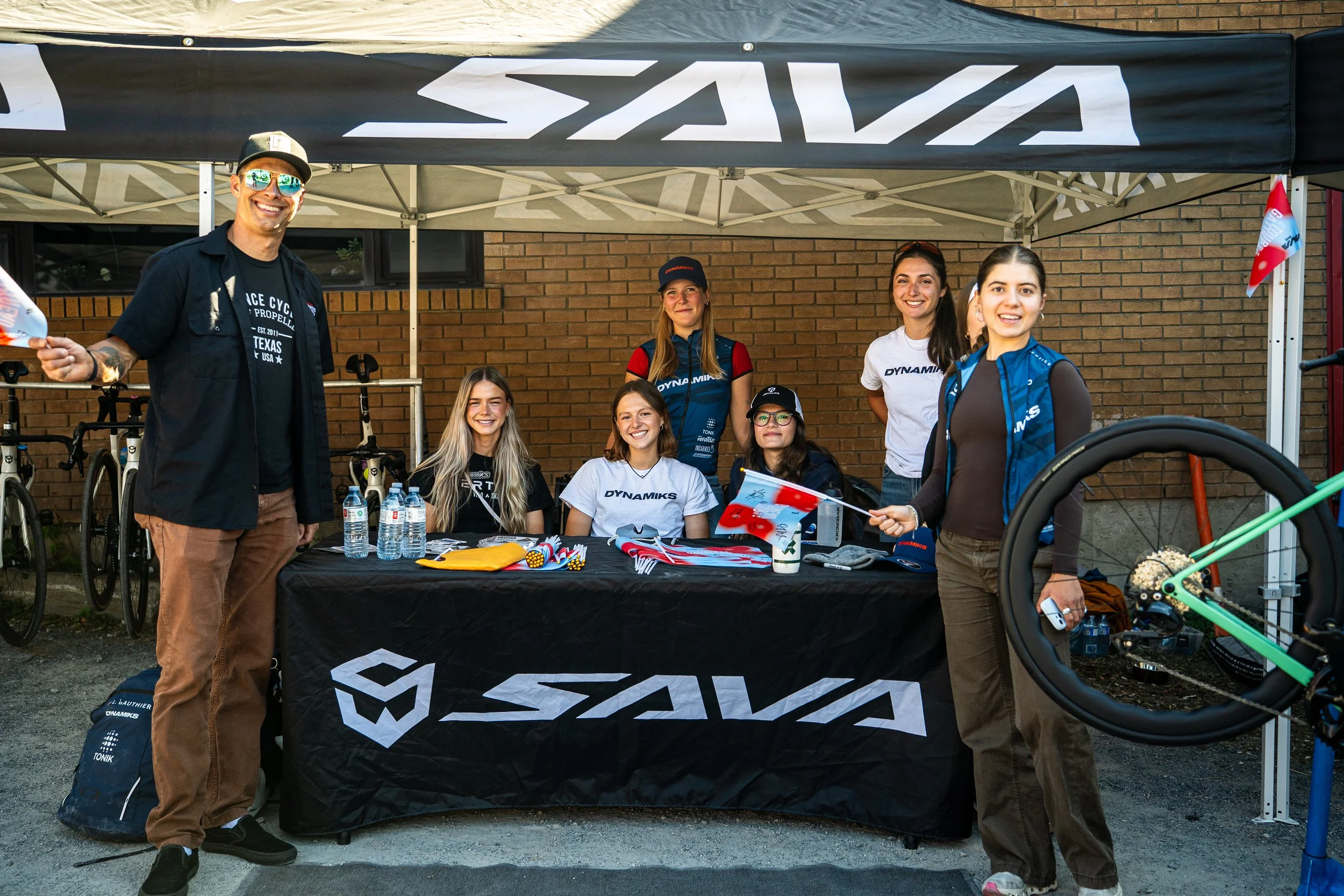 Group of people at a SAVAS booth outdoors, with bicycles in the background. Six women and one man smiling, some holding flags, under a black canopy with the SAVAS logo. The table is covered with a black cloth with the SAVAS logo and has water bottles