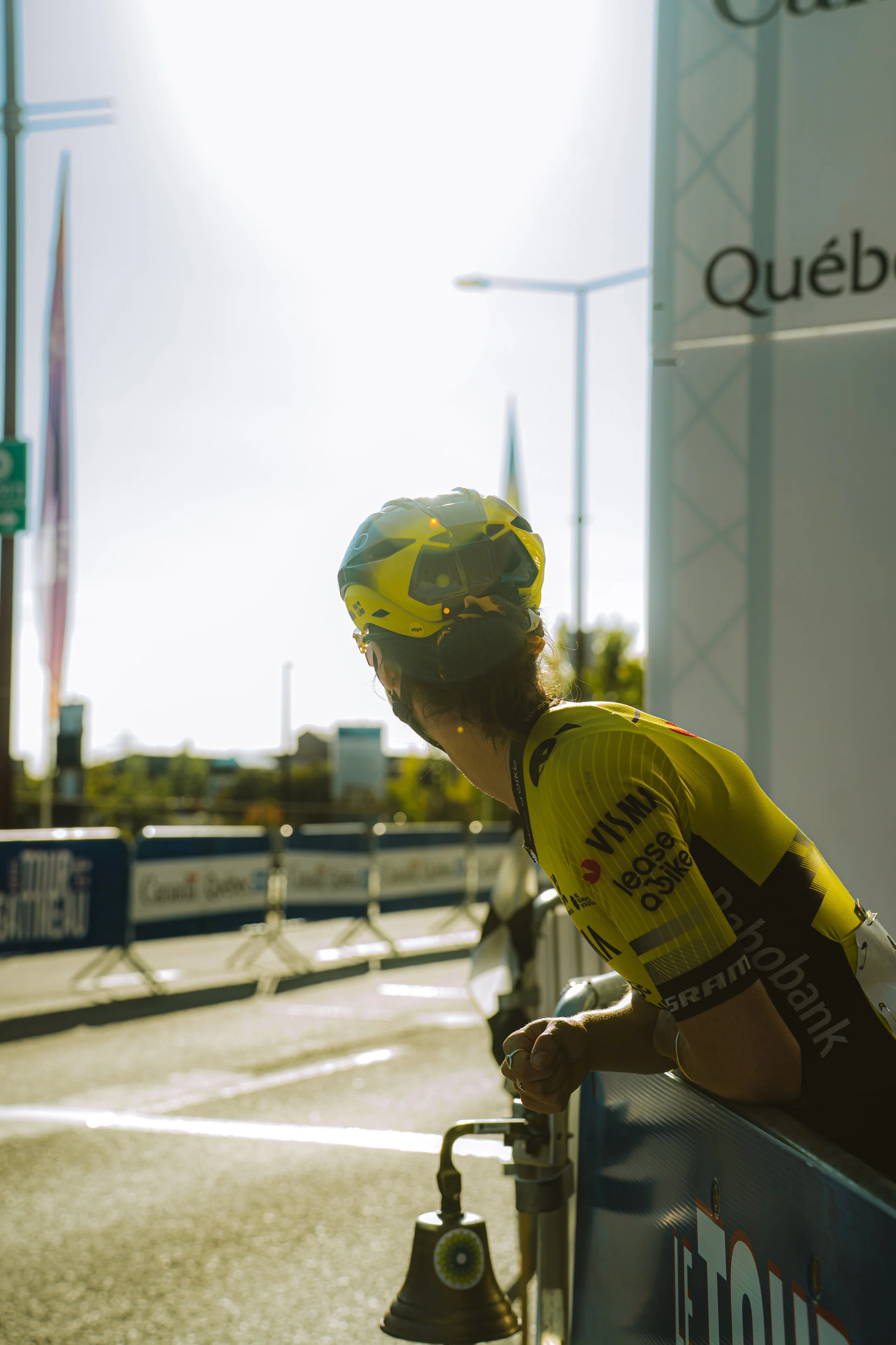 A cyclist in a yellow jersey and helmet leaning on a barricade at a race event, with the sun bright in the background.