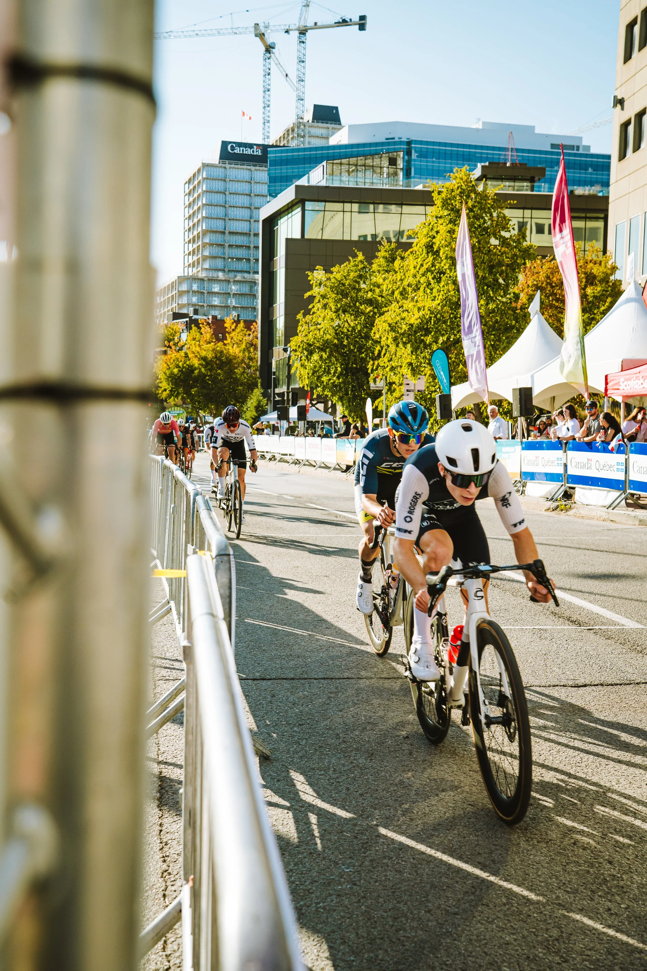 Cyclists compete in a race on a city street with tall buildings and tents in the background.