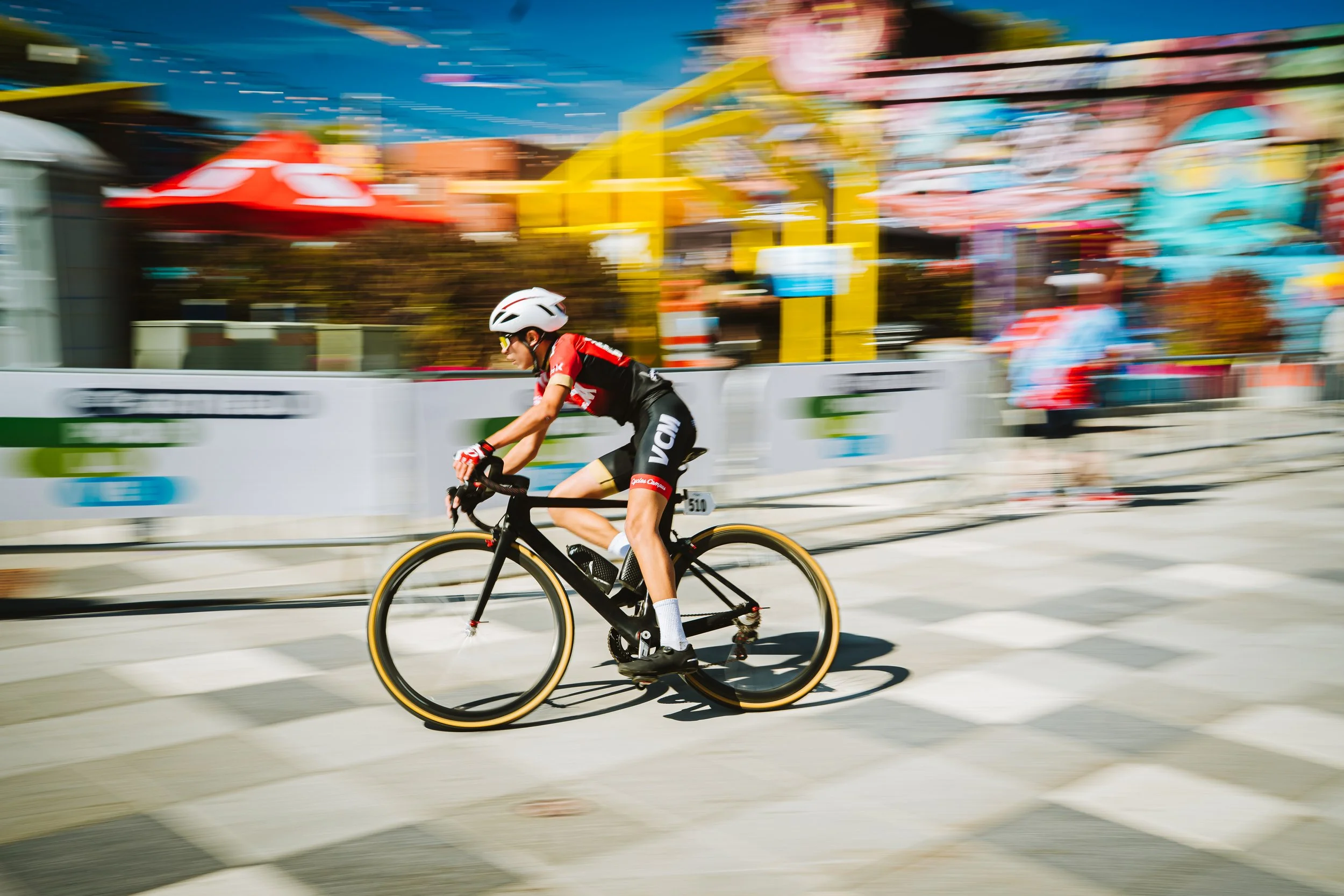 A cyclist in a black and red uniform riding a black bike during a race with a colorful amusement park in the background.