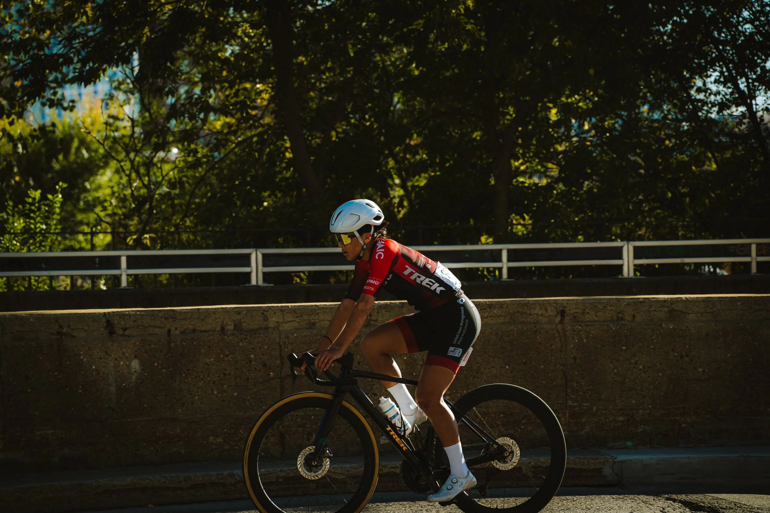A cyclist wearing a white helmet, red and black cycling kit, and sunglasses riding a black Trek road bike on a sunny day with trees in the background.