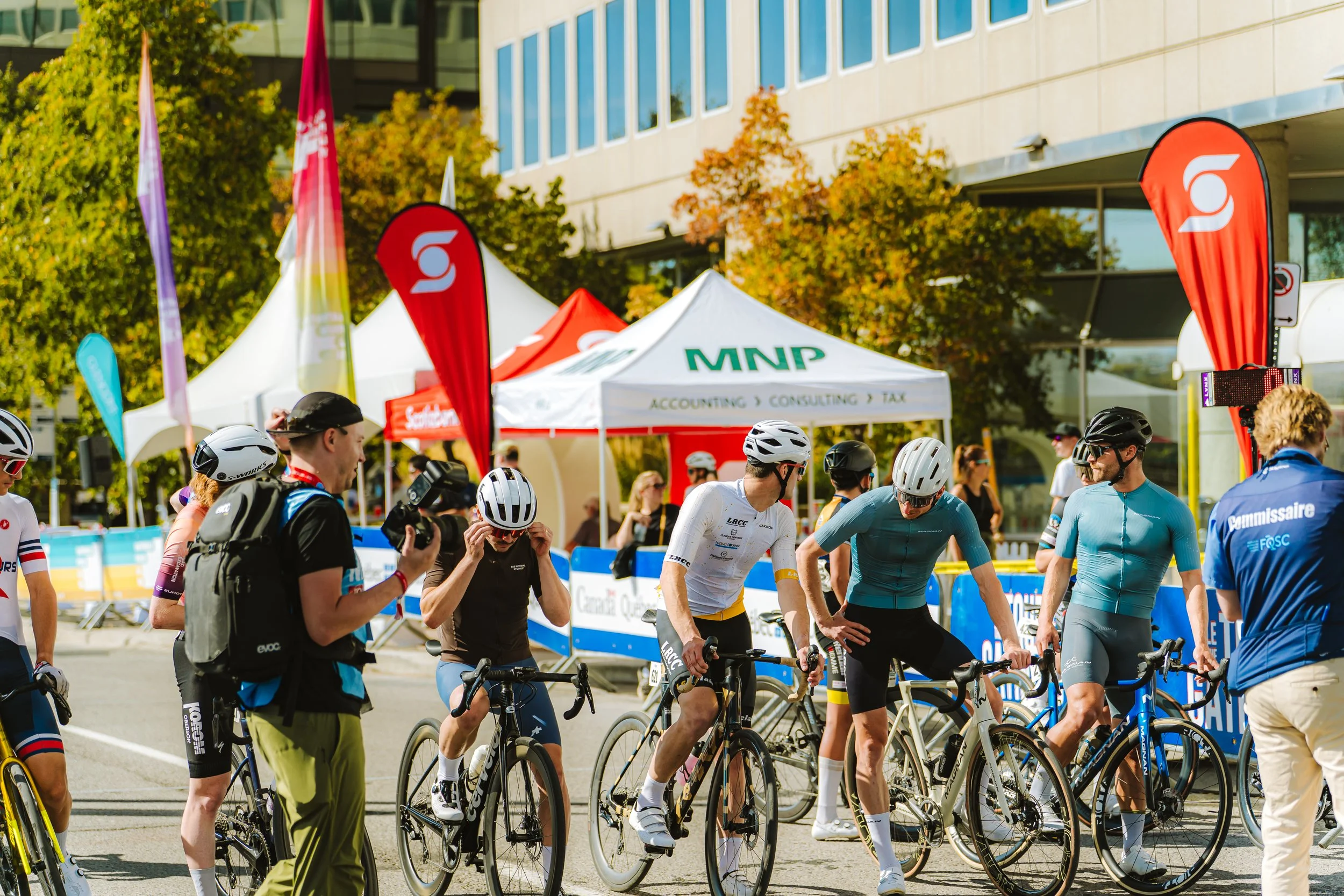 Cyclists gather with their bikes at a sports event on a sunny day, surrounded by tents and flags.