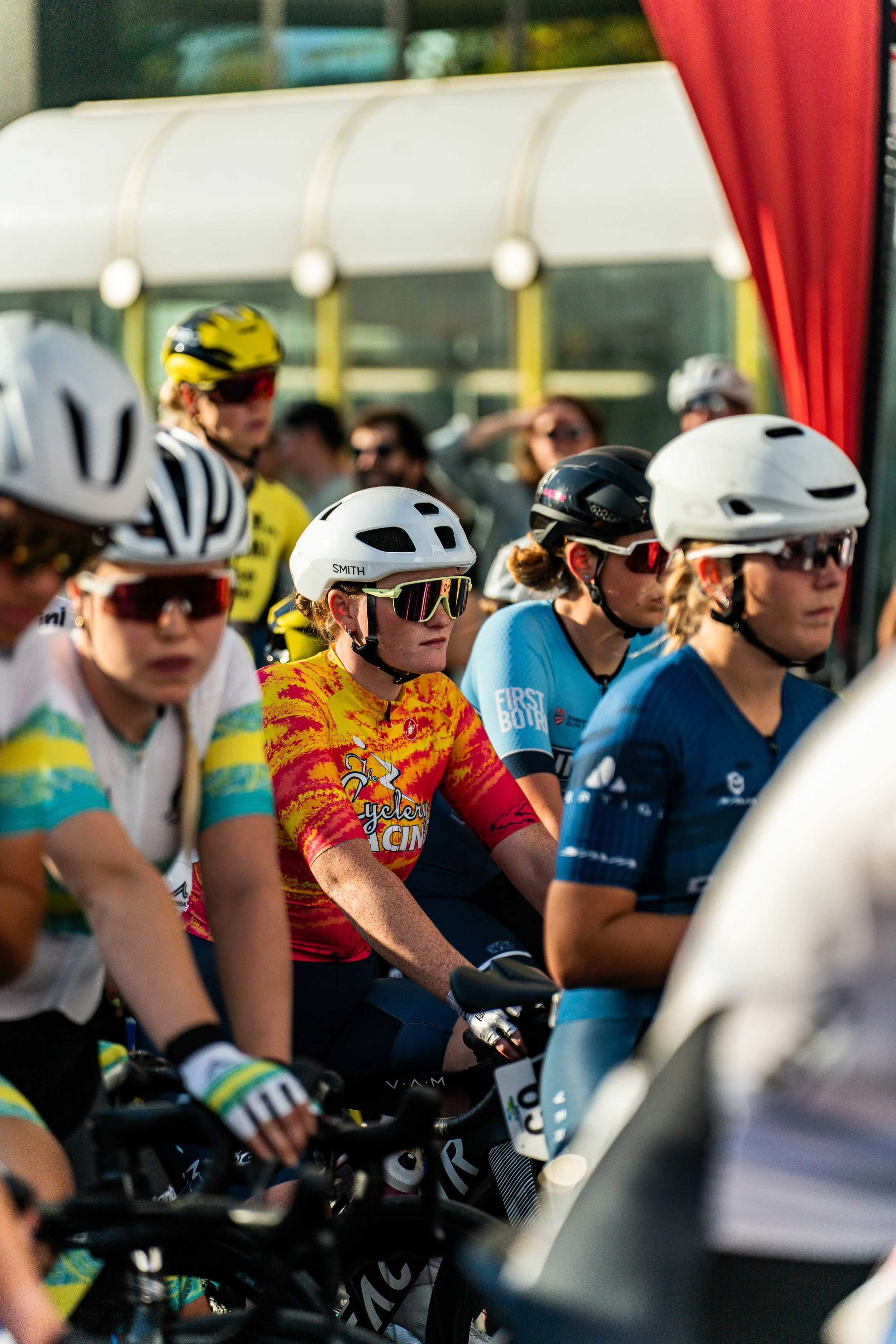 Female cyclists in colorful jerseys and helmets at the starting line of a race, with a crowd of spectators in the background.