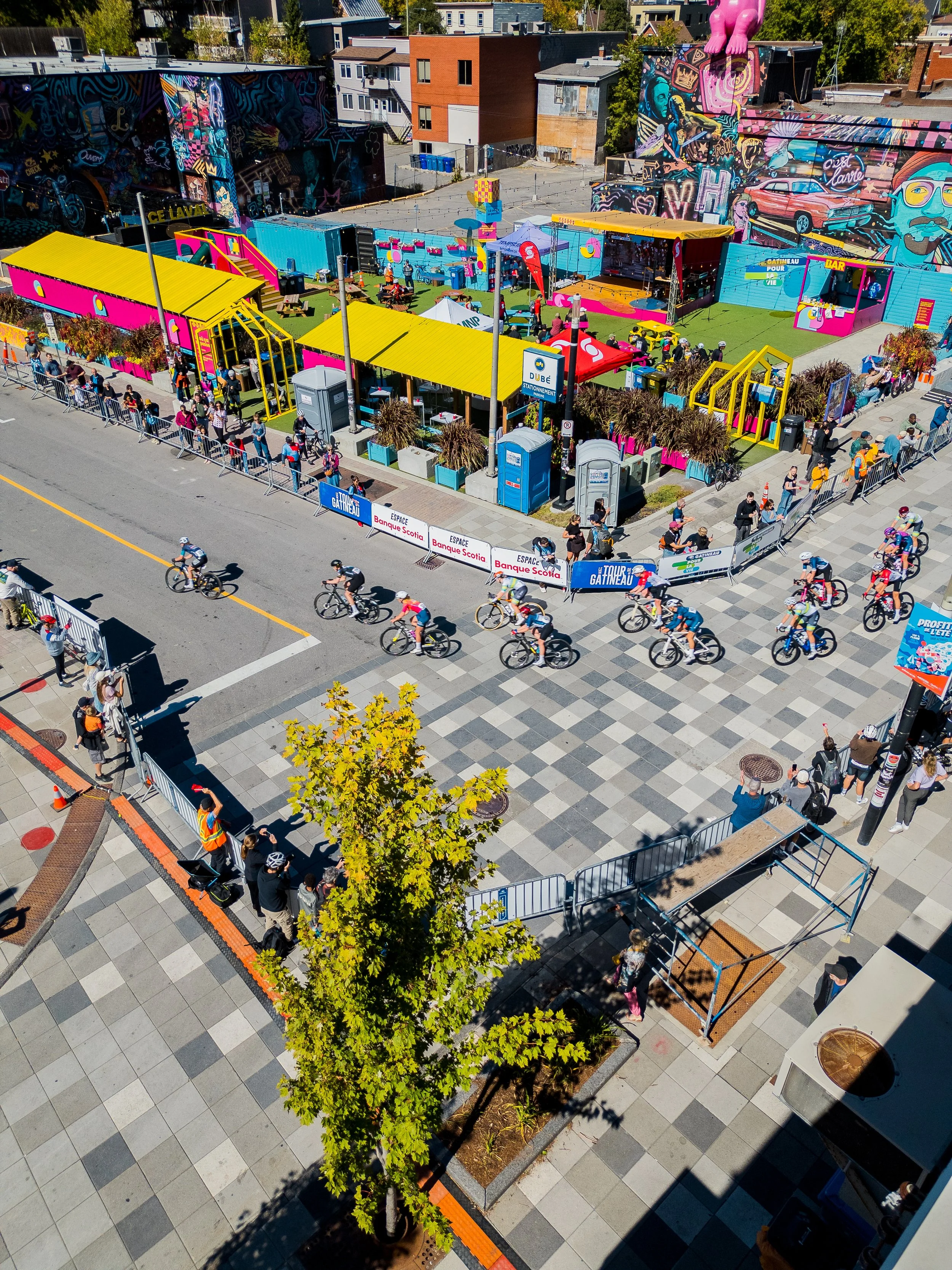 Bird's eye view of a cycling race and a colorful outdoor event with a stage, tents, and people, surrounded by murals on a city street.