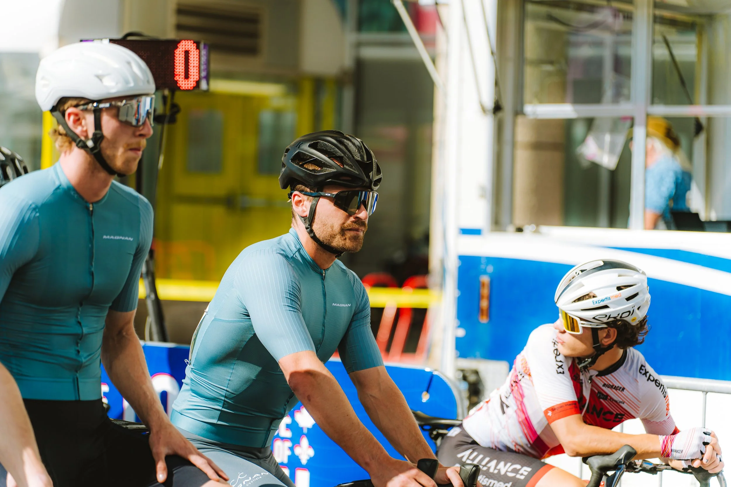 Cyclists in racing gear, including helmets and sunglasses, lining up at a race event.