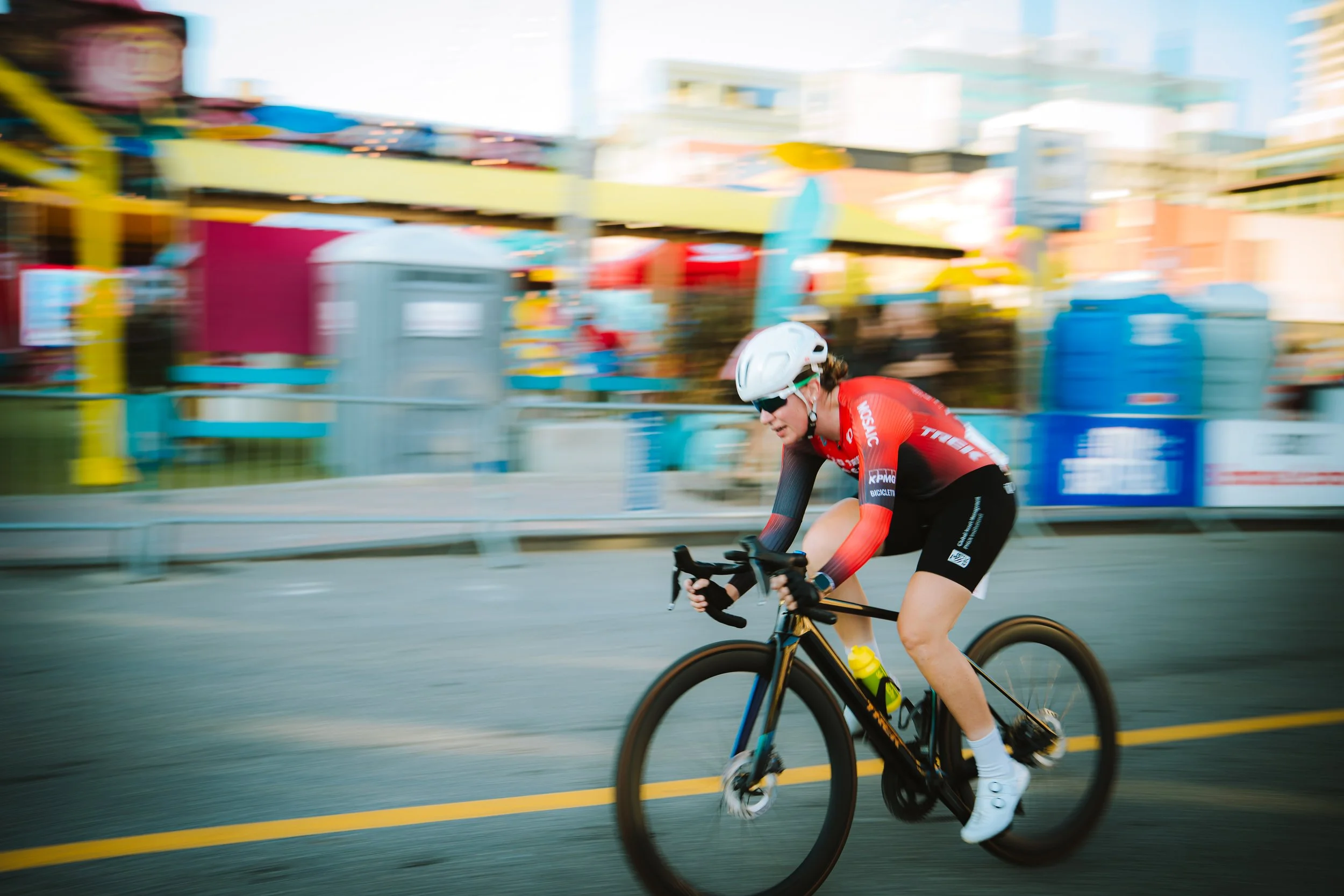 A cyclist wearing a red and black outfit, white helmet, and sunglasses riding a black bike on a city street with colorful blurred background.