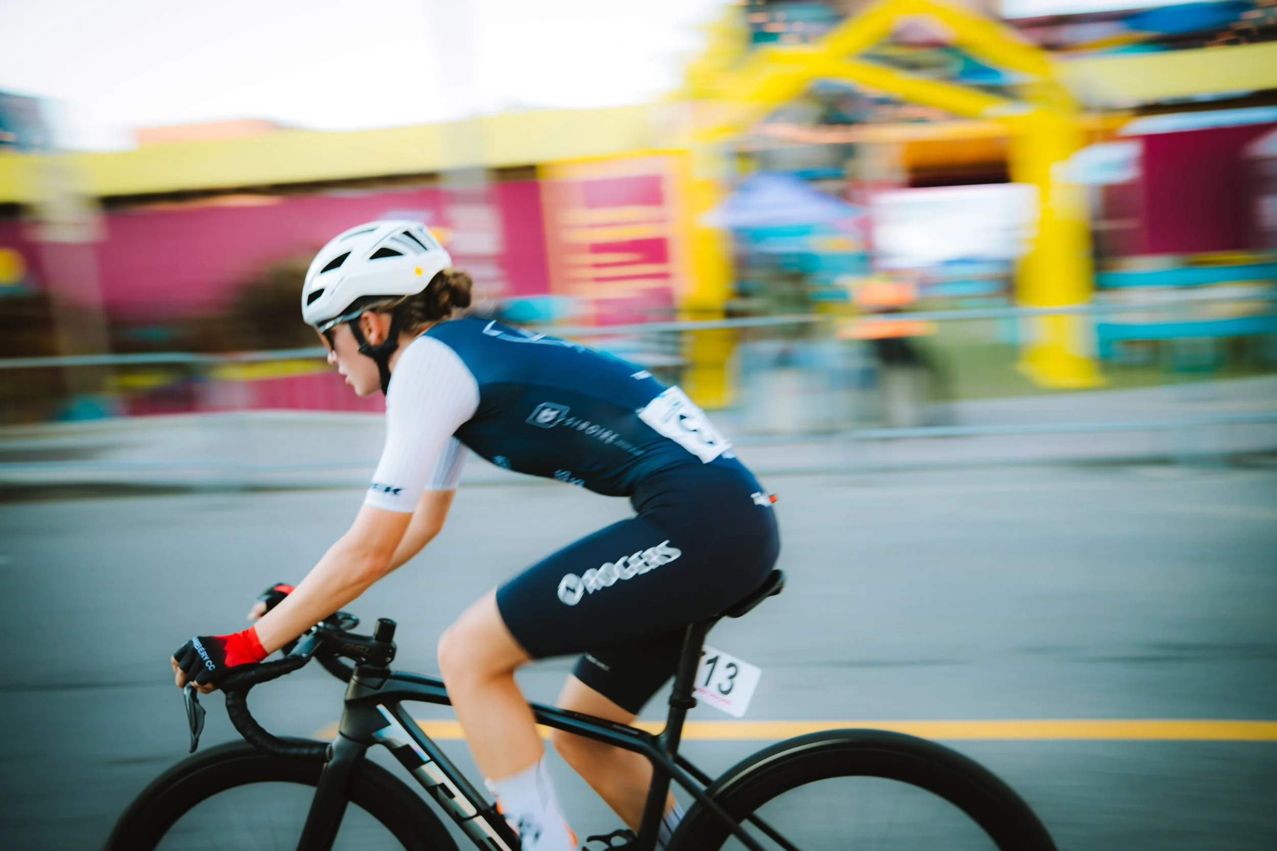 A woman wearing a white helmet, navy blue cycling jersey, black shorts, and red gloves riding a black road bike during a race, with a blurred colorful background.