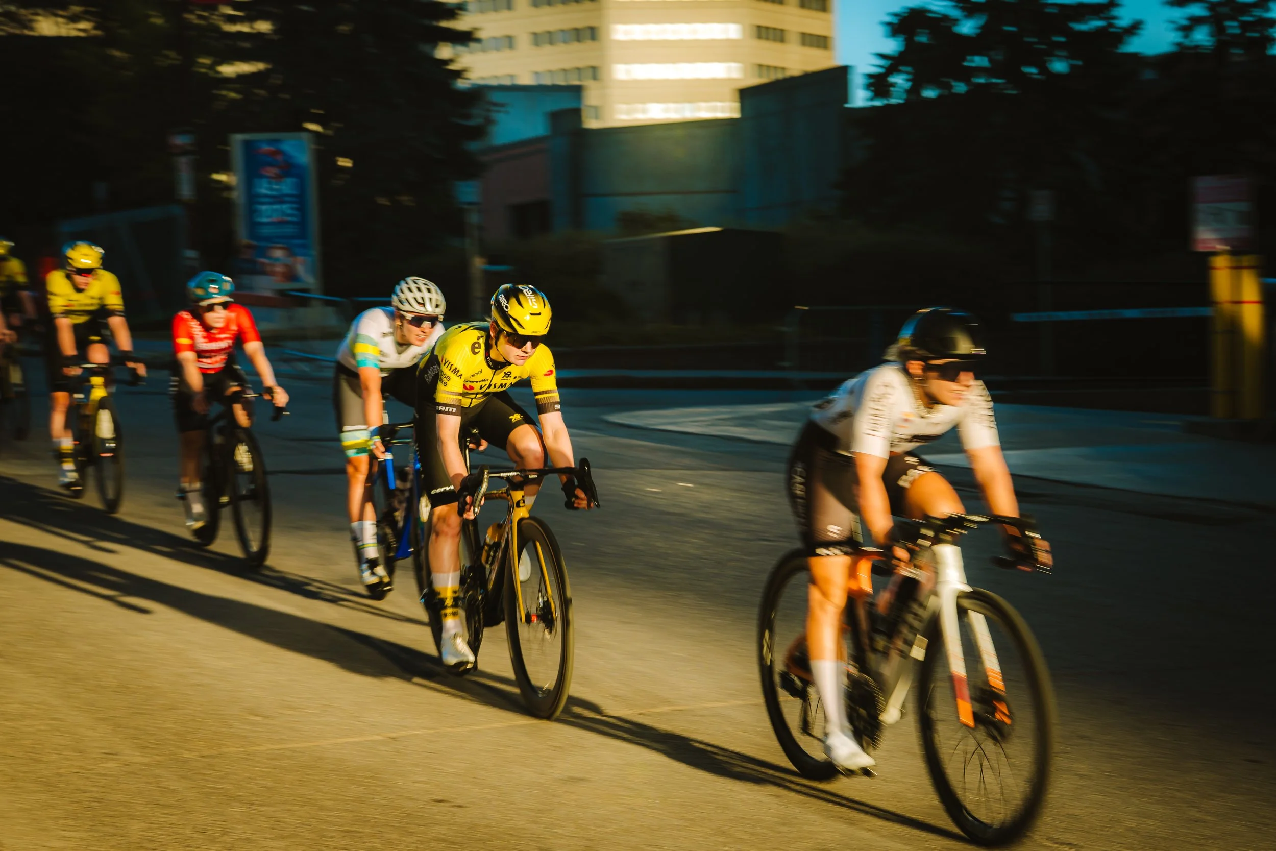 Group of cyclists racing on a city street at dusk, wearing colorful jerseys and helmets.