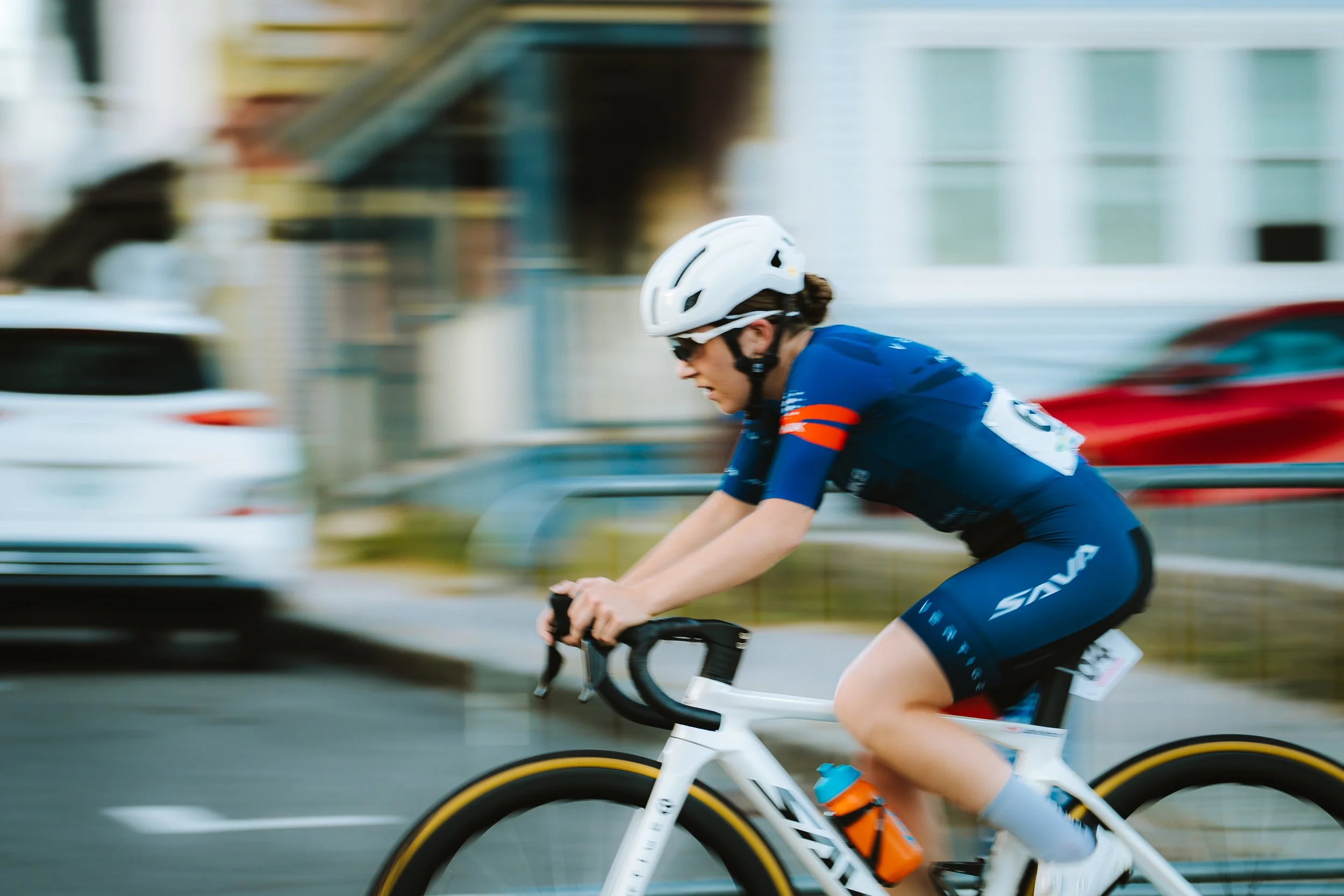 A cyclist wearing a blue jersey, black shorts, white helmet, and sunglasses riding a white racing bike at speed on a city street during daytime.