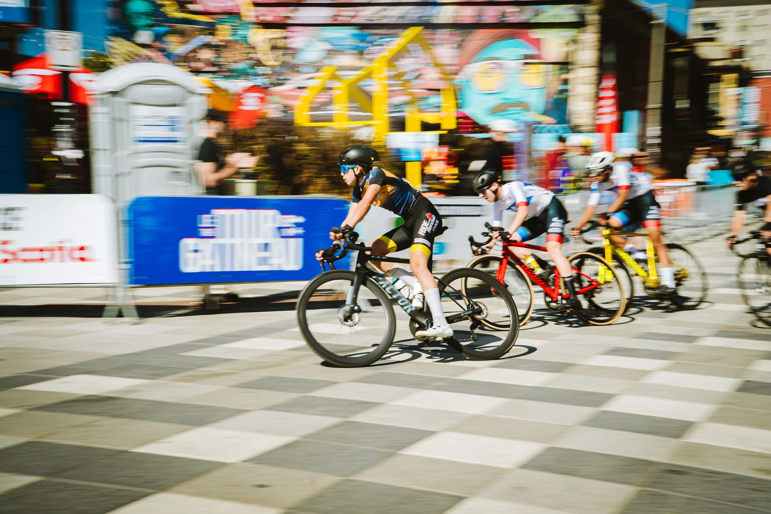 Cyclists racing on city streets during a bike race with colorful buildings and banners in the background.